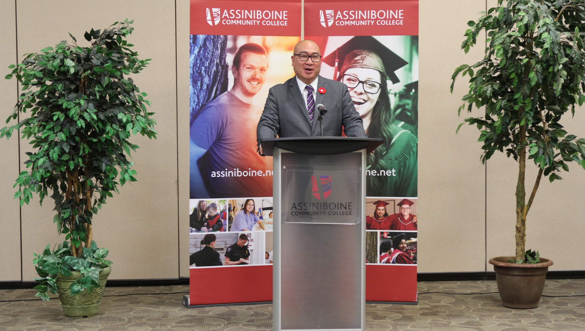 The Honourable Jon Reyes, Minister of Advanced Education, Skills and Immigration speaks at an Assiniboine Community College podium with college banners behind him. 