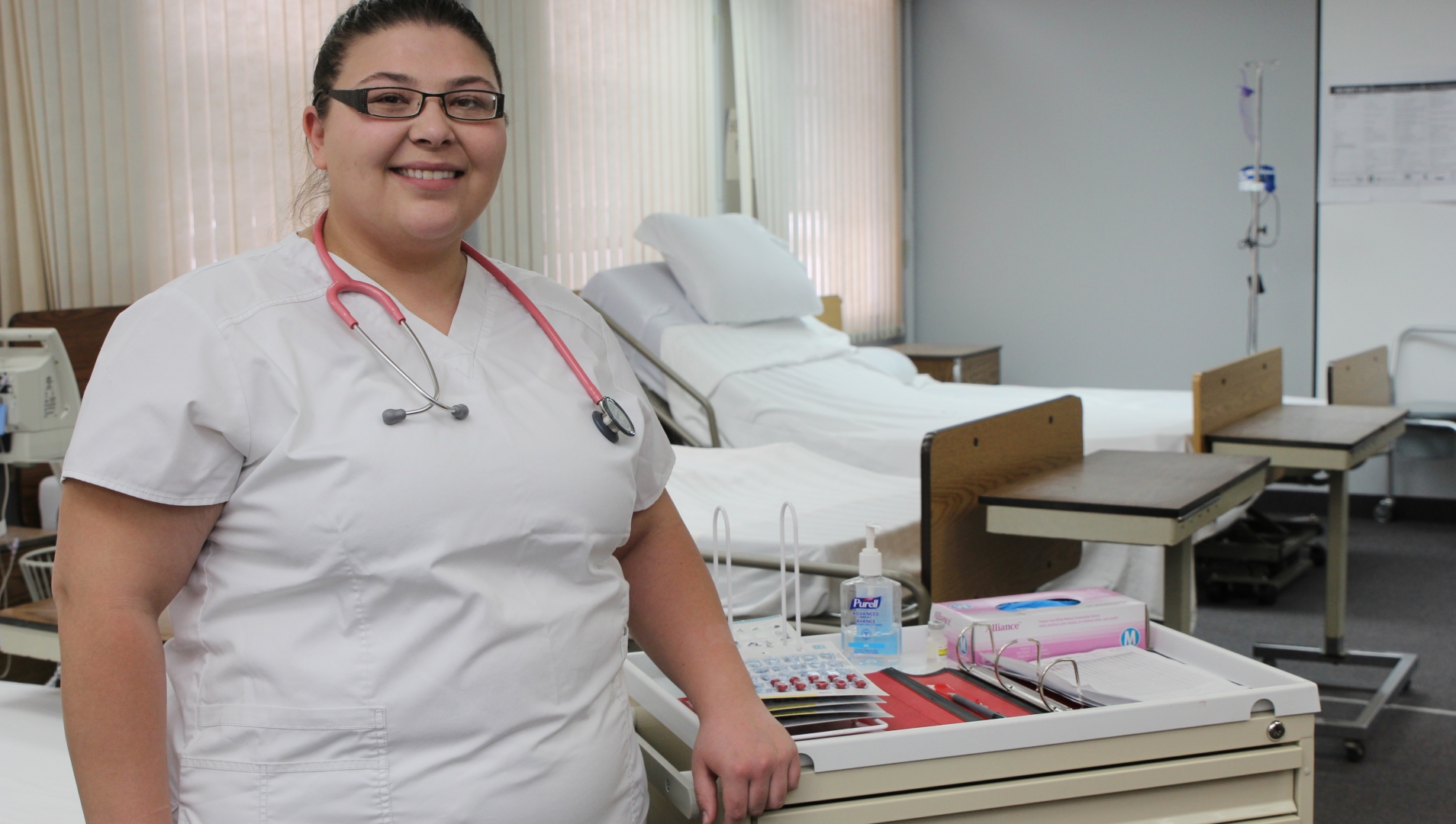 A nursing student stand in the foreground, in the left of the frame. Behind her are training beds and equipment.