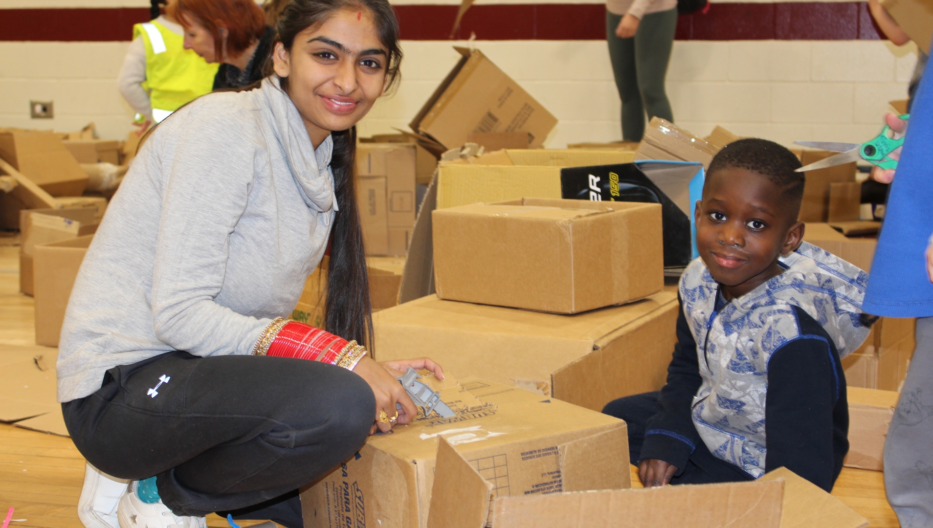 A woman in the left of the frame crouches down beside a young boy who appears in the right of the frame. They both smile at the camera, surrounded by empty cardboard boxes they appear to have been playing with. 