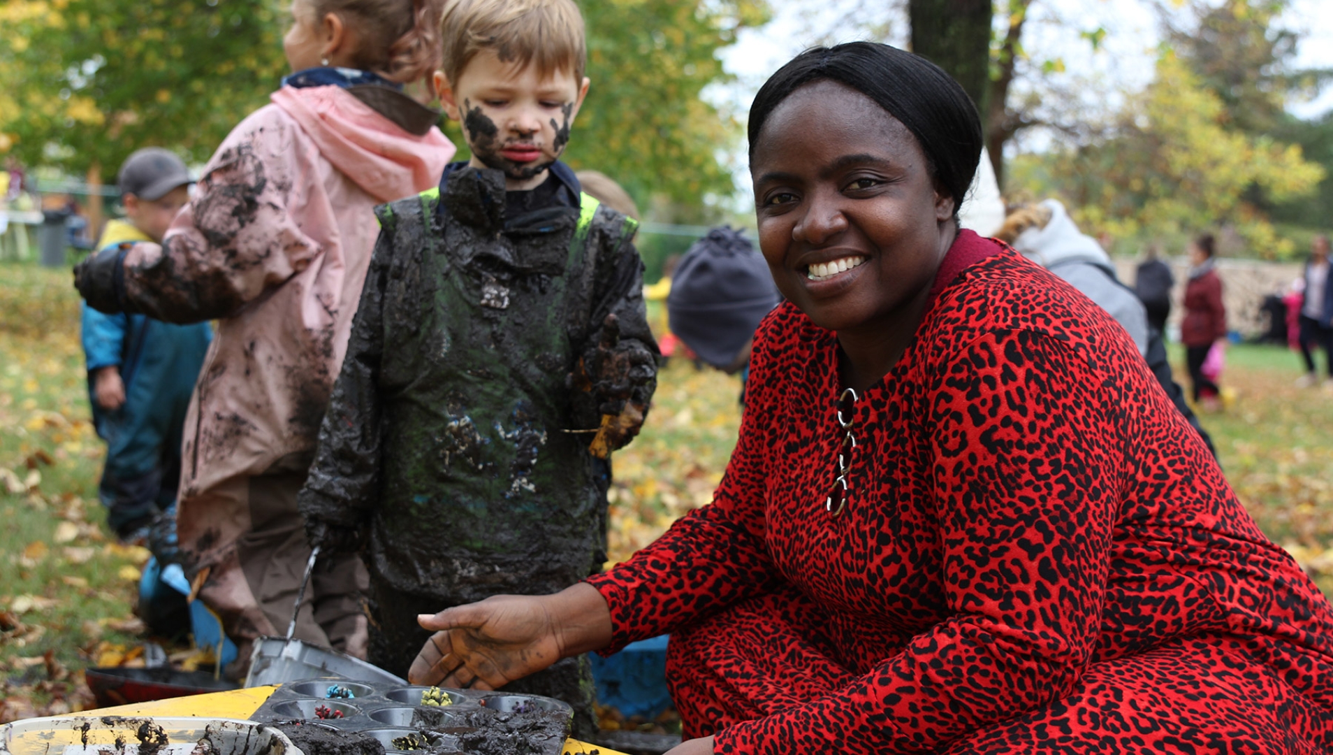 A smiling person in a red-and-black dress with a mud-covered child standing next to them and more children running in the background.