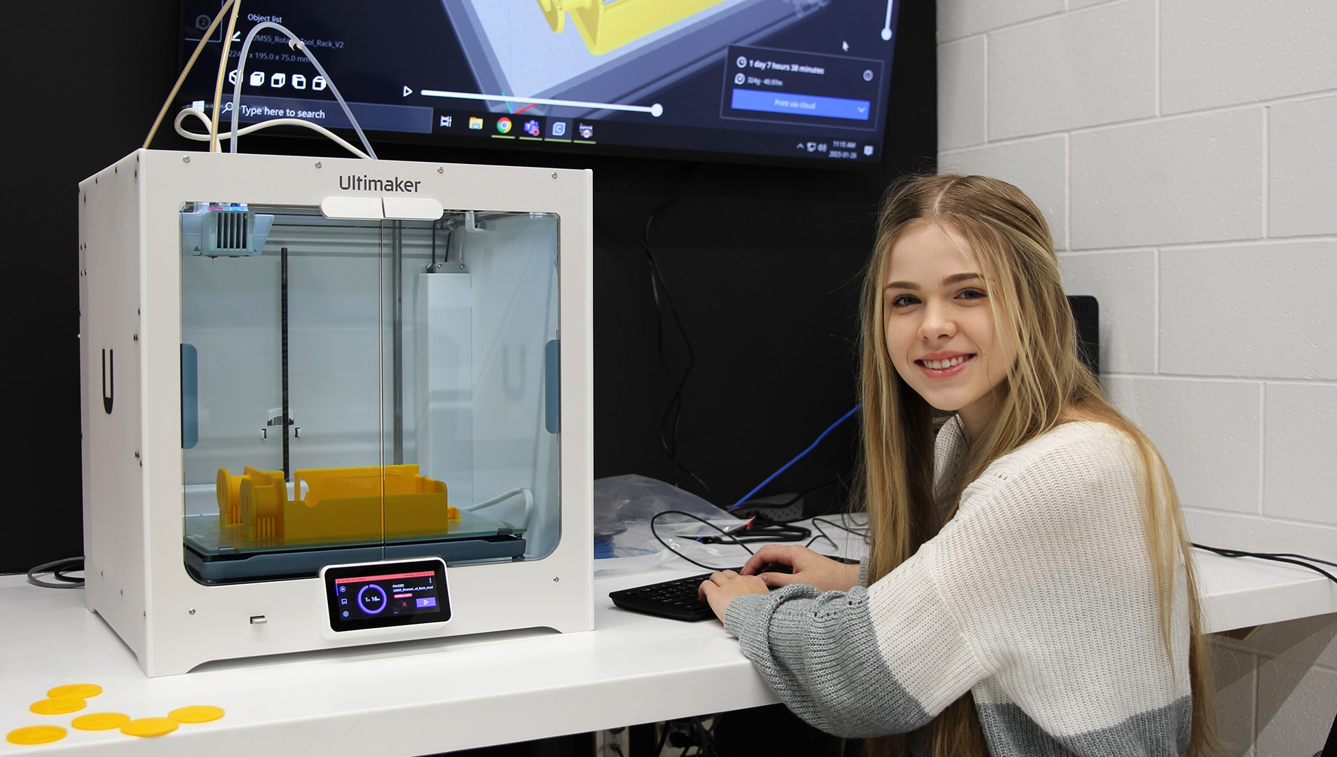 A female student sitting in front of a 3D printer with a big display above it, inputting printing instructions.