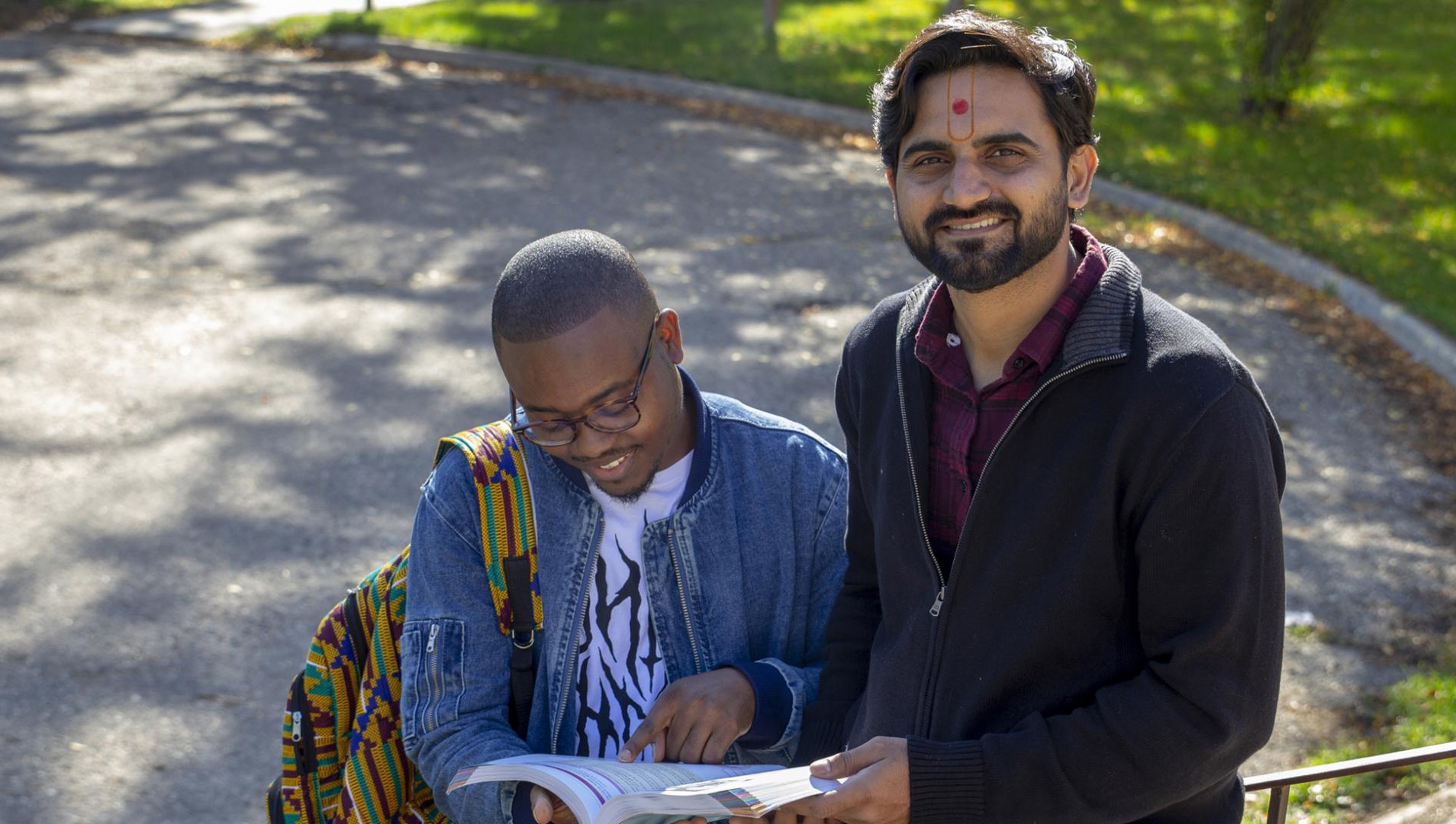 Two male students standing outdoors with a green lawn full of fallen leaves behind them, looking at a textbook.