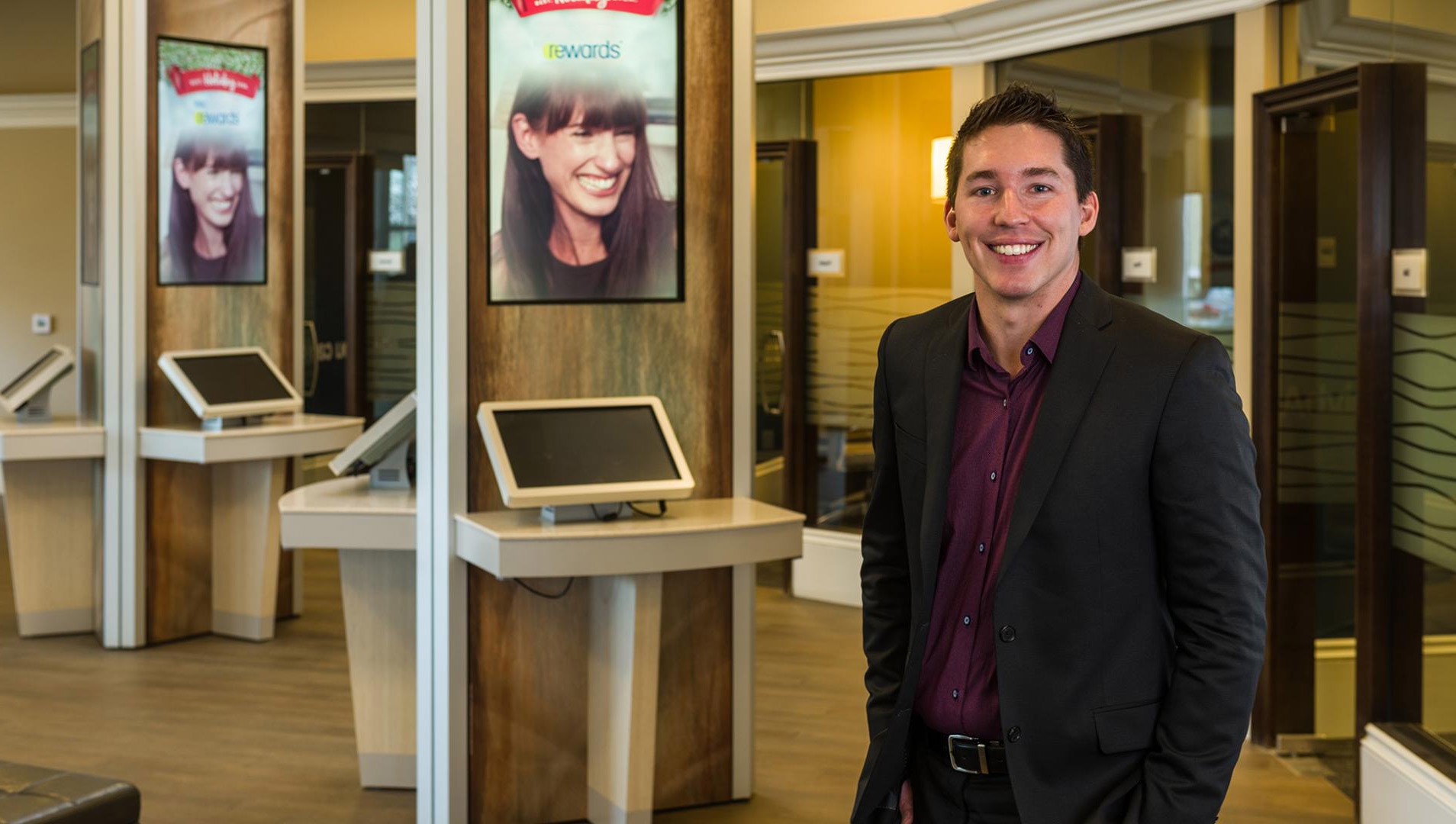 A man in a black suit with a burgundy shirt standing in the middle of an empty customer area in a bank, smiling.