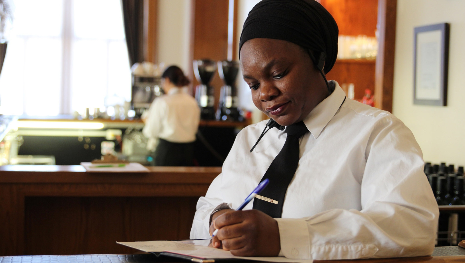 A hospitality business management student in a white shirt and a tie writing notes while standing behind the counter.