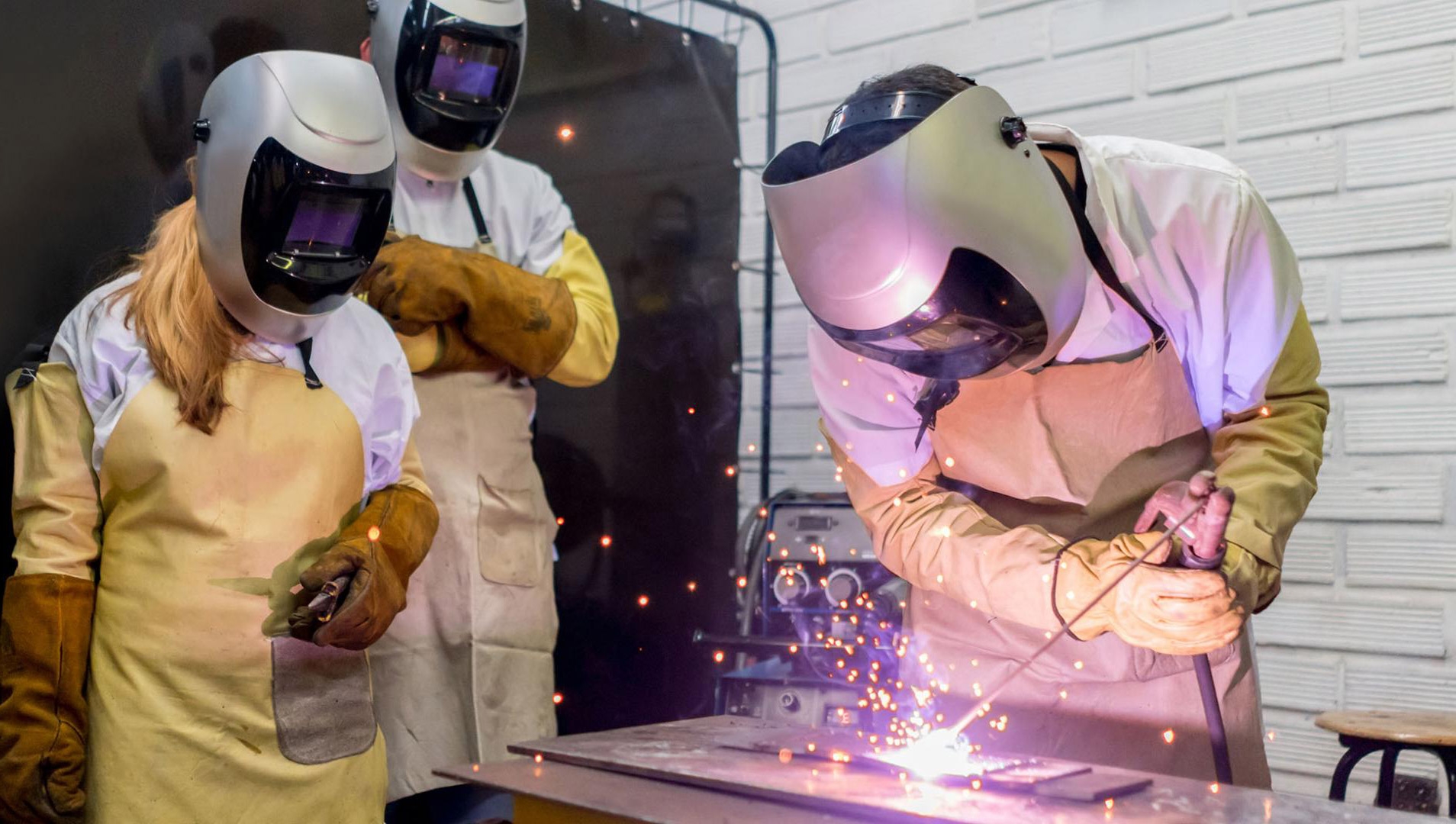 Three students in welding uniforms and helmets, one of them is welding metal pieces together on a surface in front of them.