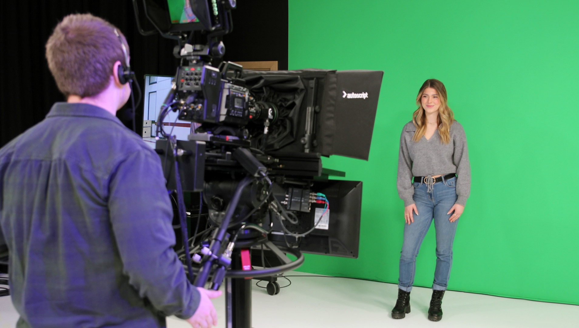Two media and communications students, one standing behind a professional TV camera, recording the other student in front of a green screen.