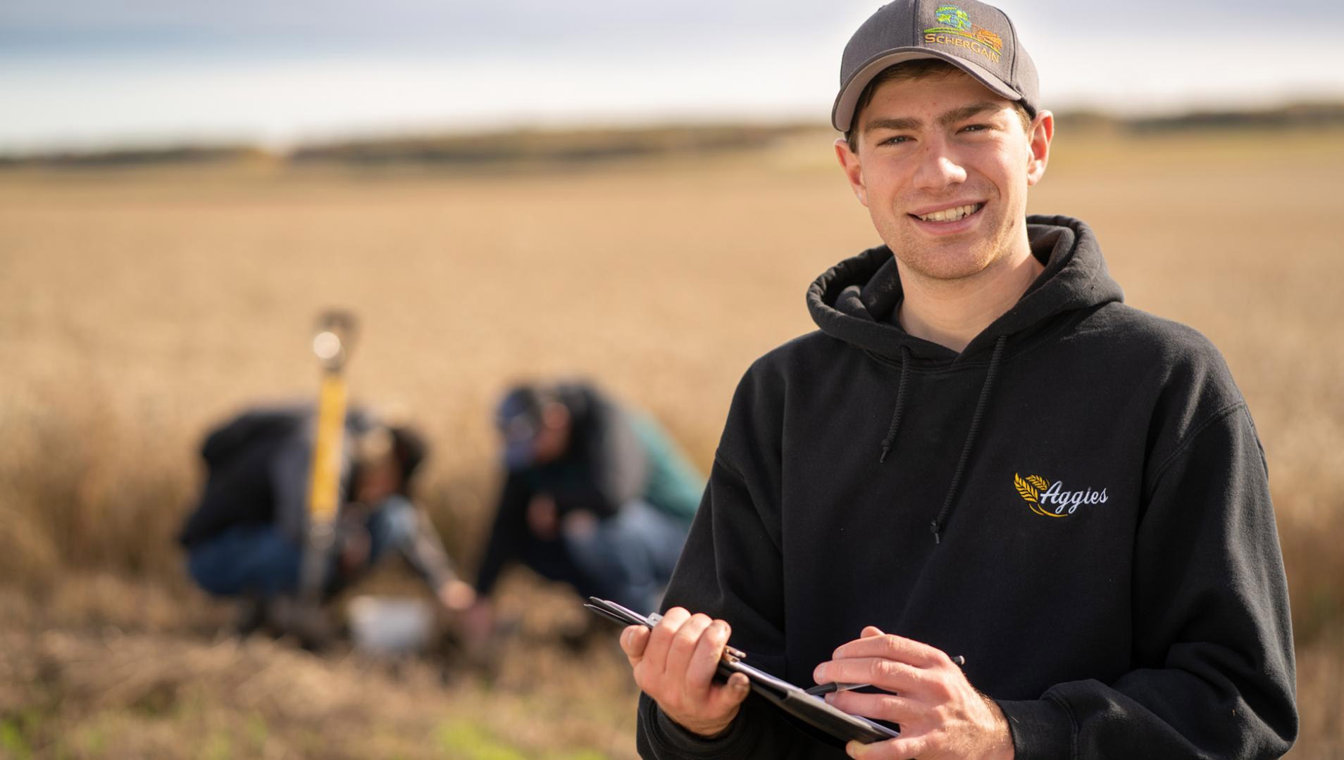 On the right side of the picture a male agribusiness student wearing a grey cap and black hoodie is holding a clipboard and smiling at the camera. In the background there is a wheat field and two other students are looking at something on the ground.