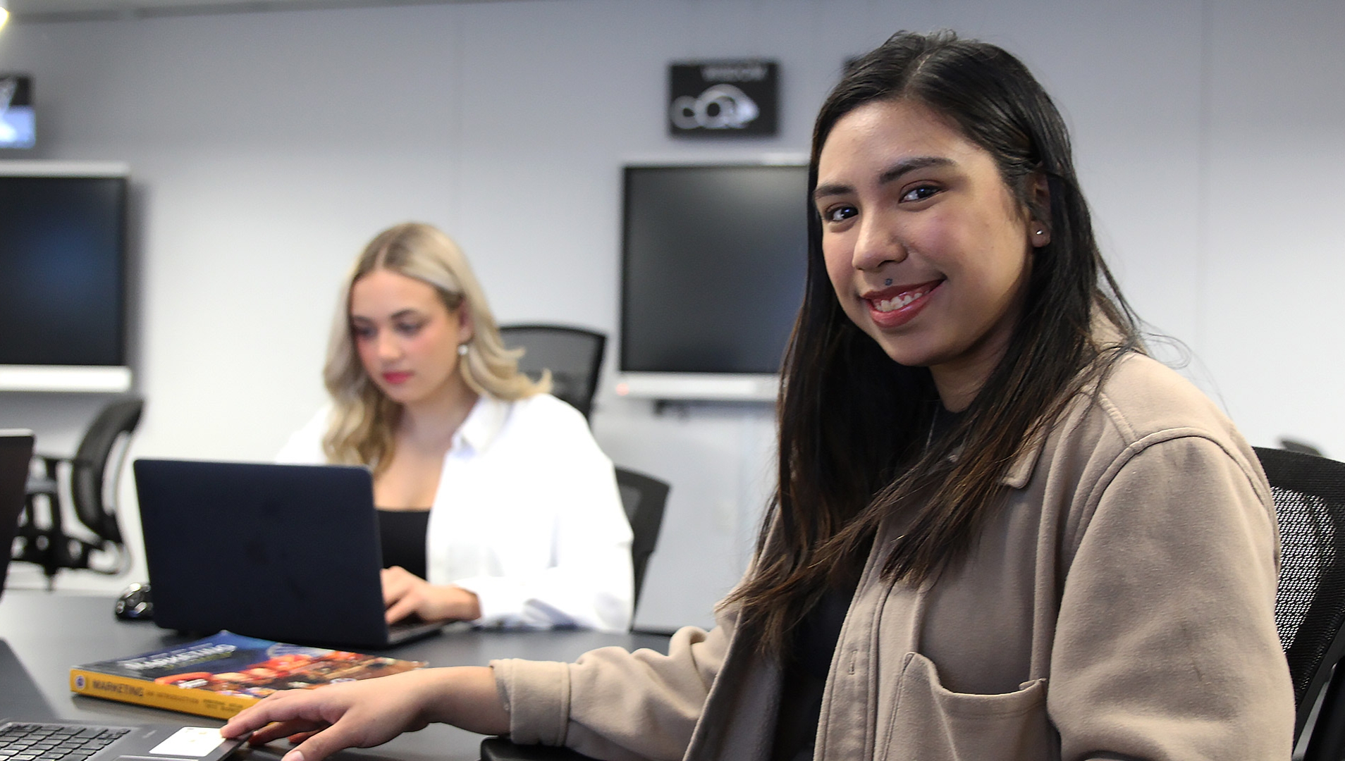 On the right side of the picture there's a female indigenous student sitting at a desk in the Westoba Lab room with a laptop and a textbook, smiling at the camera. In the background, on the left side of the picture another female student is using a laptop.