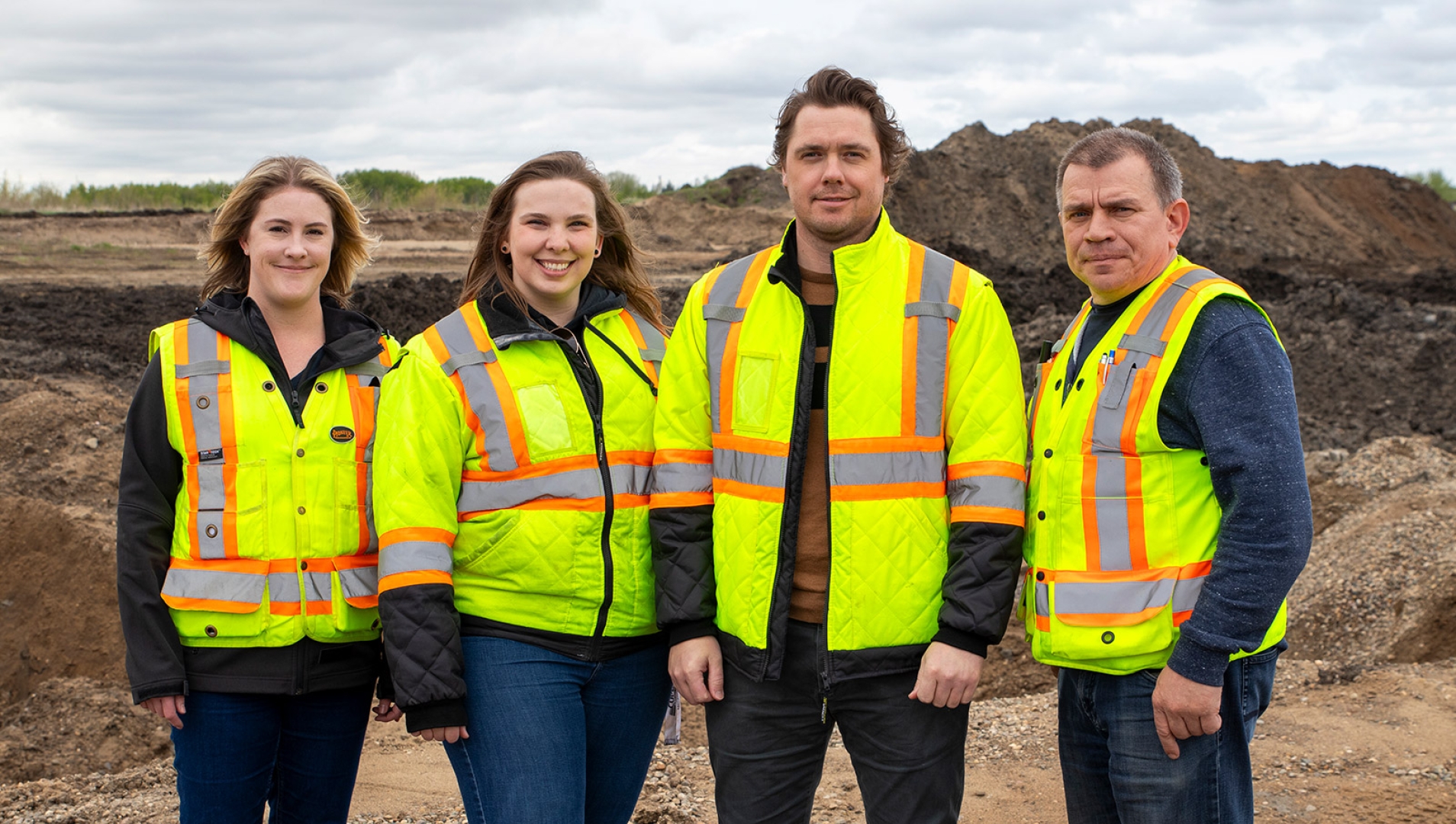 Four civil technician students in reflective vests standing together with mountains of dirt behind them.