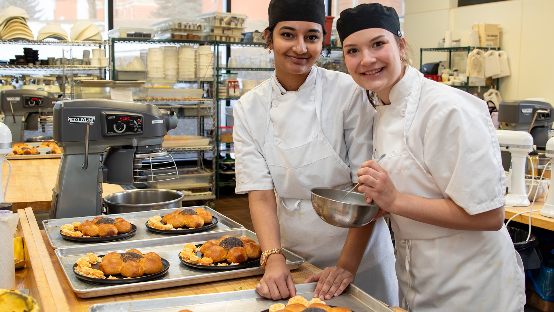 Two female students in kitchen uniforms standing in front of a counter full of pastries.
