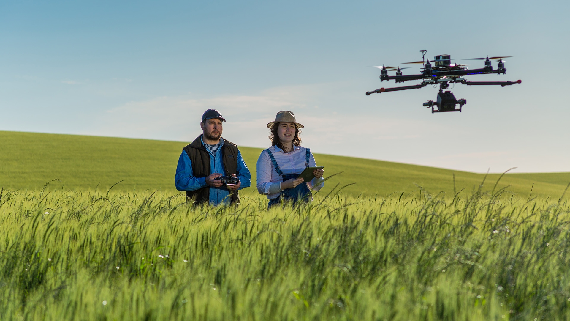 Two people standing in a field, one of them is operating a drone flying in front of them.
