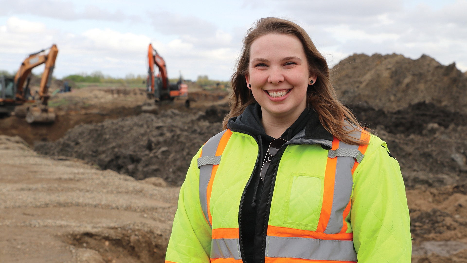A female civil technician student in a yellow high-visibility jacket standing at an excavation site.