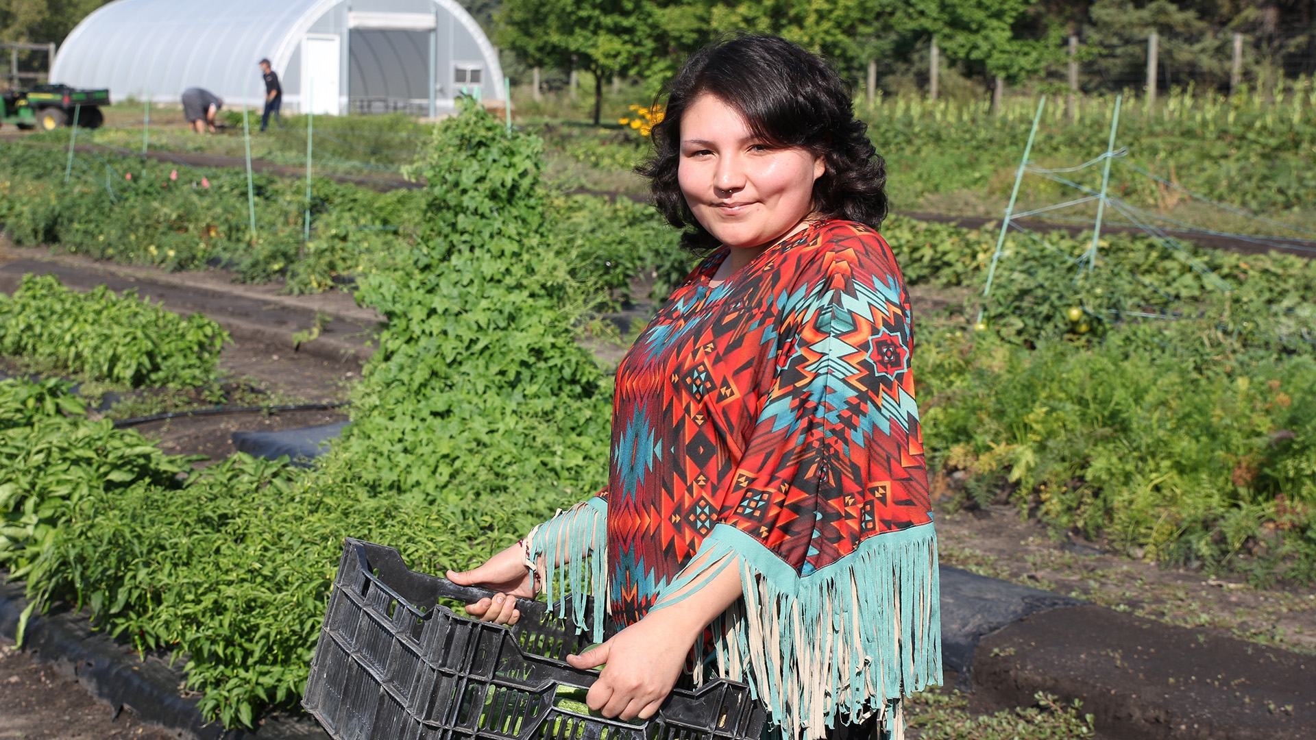 A student in a blue and red patterned poncho holding a case of plants, standing between farming plots.