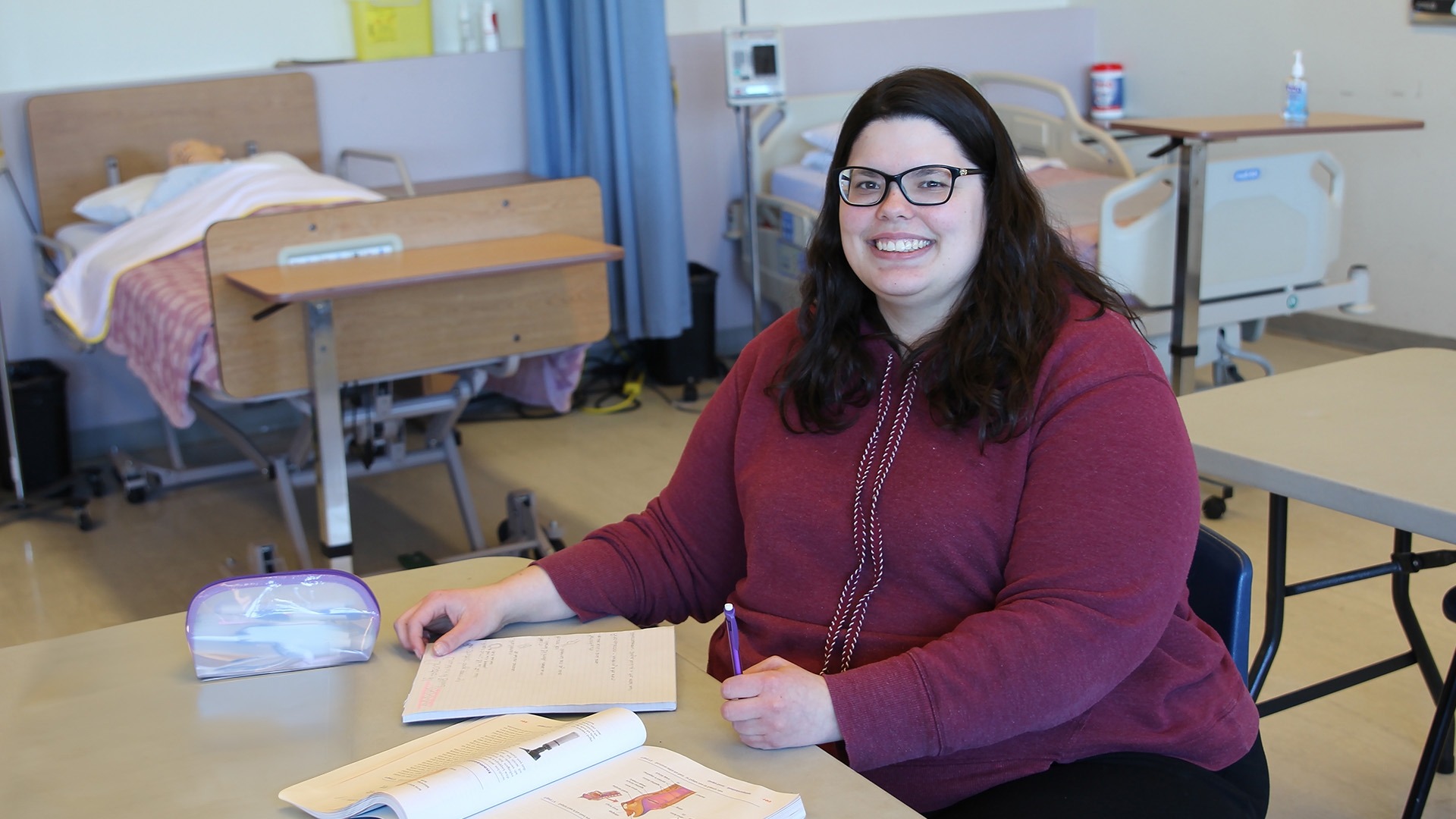 A medical administration student sitting at a desk in a nursing classroom with textbooks in front of her.