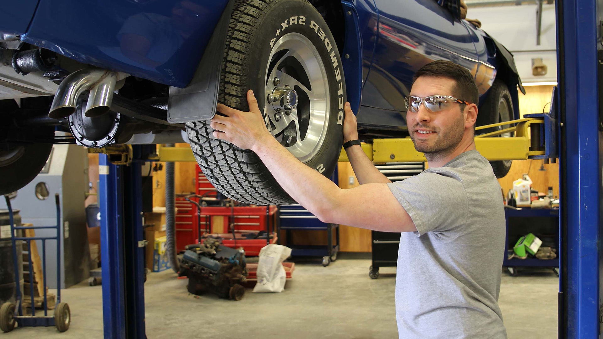 Automotive technician standing in front of a blue sports car on a lift, changing a tire.