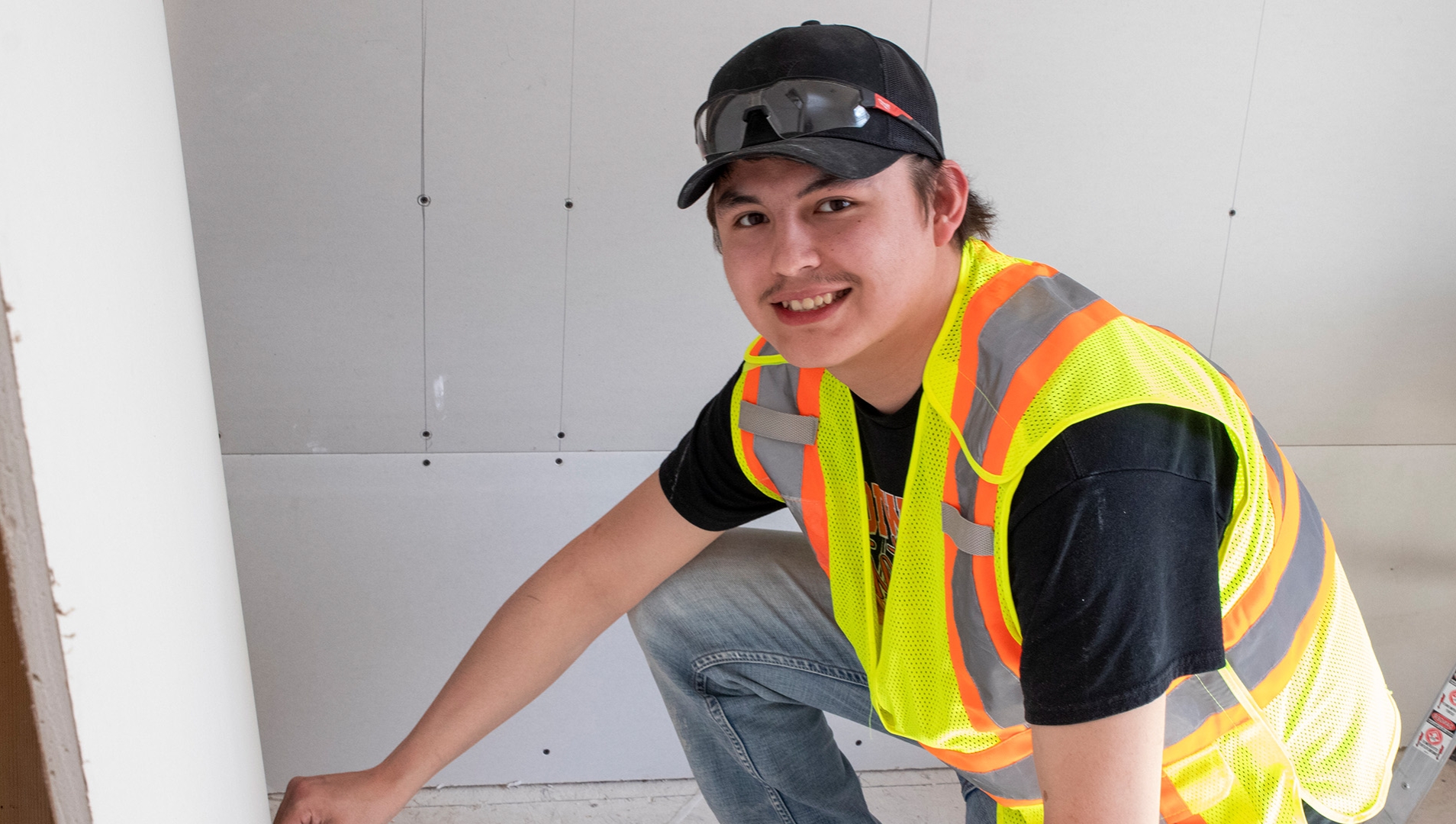 Carpentry student in a high visibility vest kneeling next to a piece of drywall.