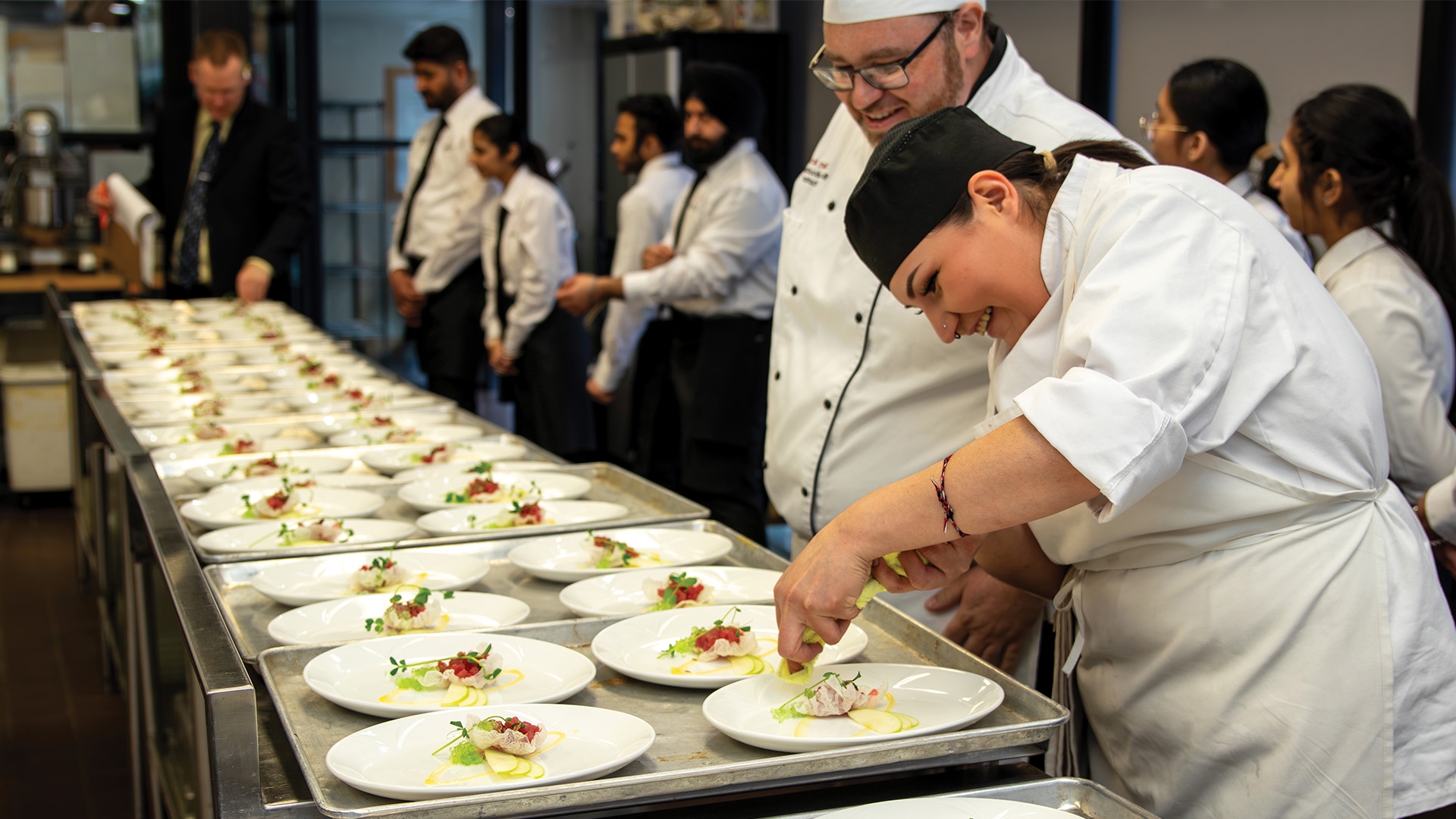 Two culinary students standing in the kitchen in front of two rows of plates with food on them, and a line of students in the background. One of them is adding finishing touches to the dish, the other one watching it, both of them smiling.
