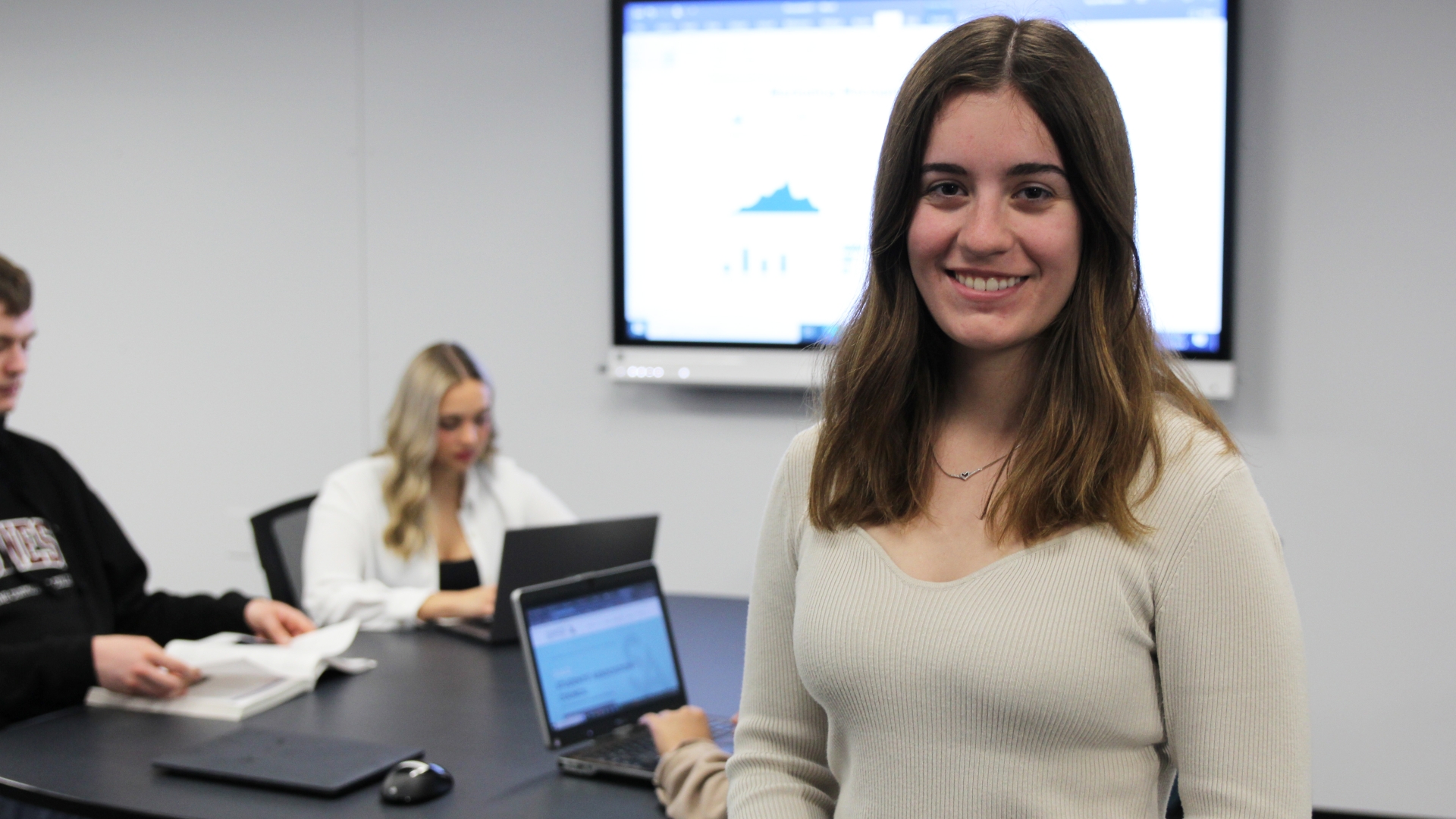 A marketing student standing in the Westoba Lab with a table full of other students behind her and a screen displaying a data report.