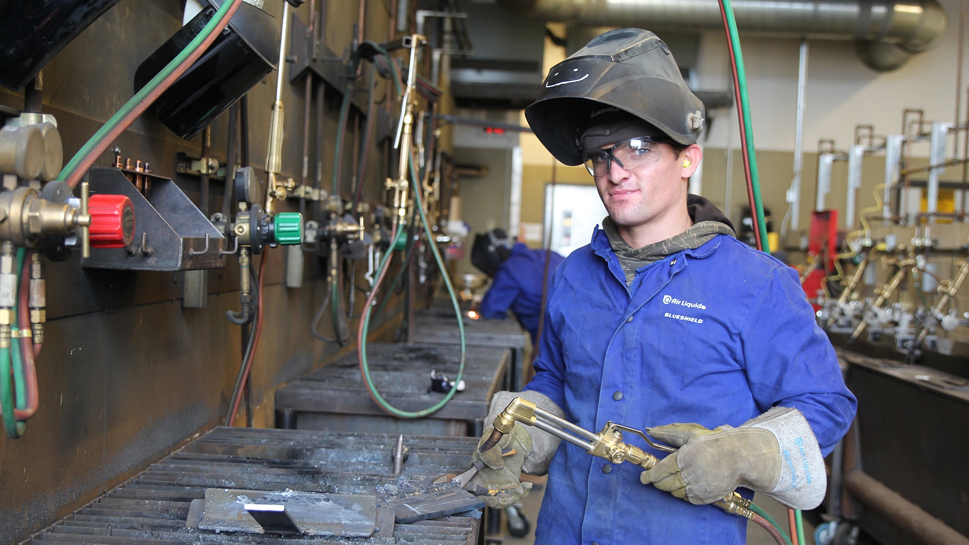 A welding student in a mask and gloves standing with a welding torch in a shop.