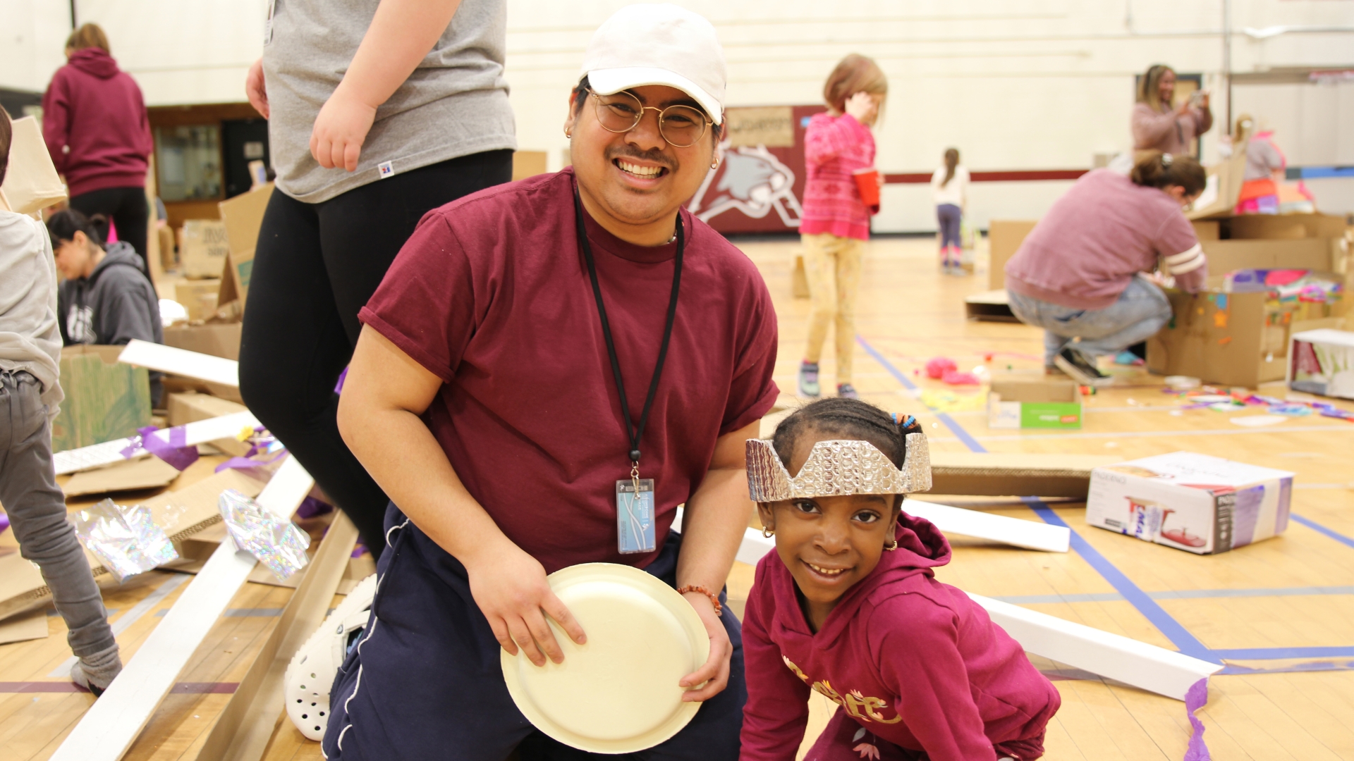 An early childhood education student kneeling next to a kid wearing a bubble wrap crown, both of them smiling, with other students and kids playing in the background.
