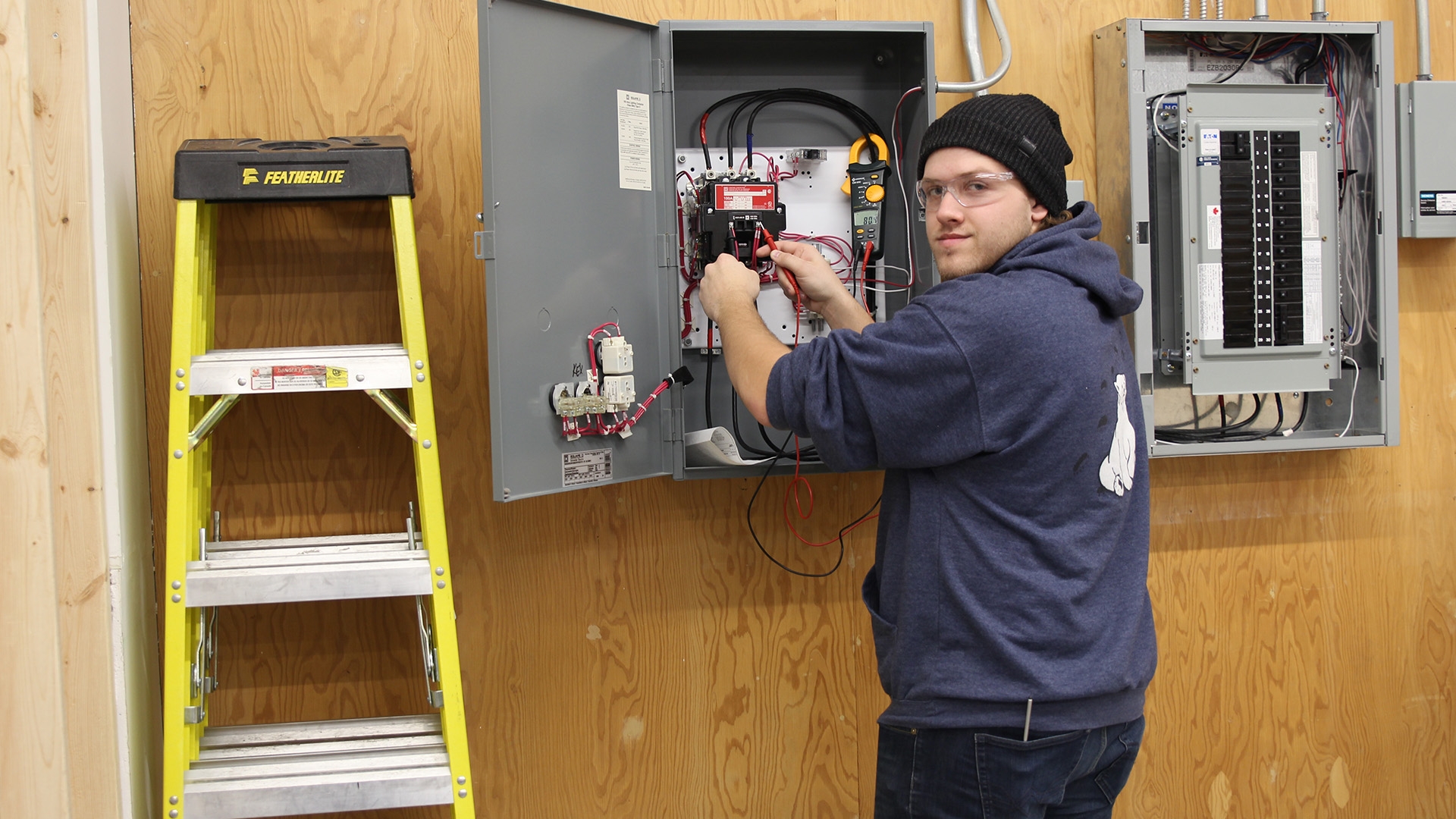 A student working on an electrical box with a ladder leaning against the wall next to it.