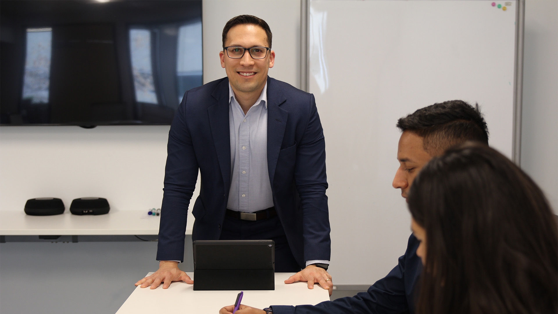 Financial Services student standing over a desk with a tablet on it, other students sitting to the right of the desk.