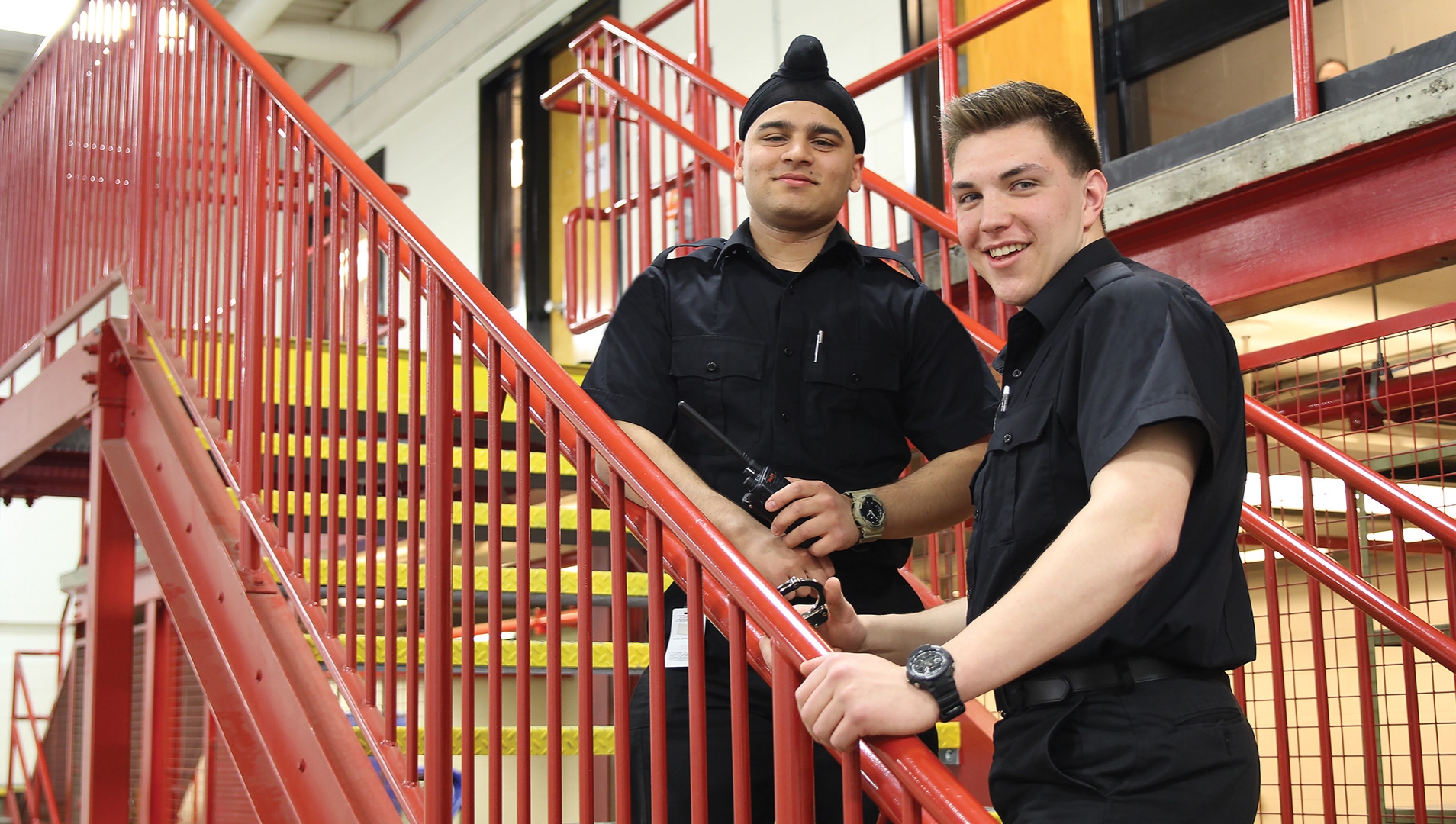 Two male public safety students in black uniforms standing on a staircase with red railing.