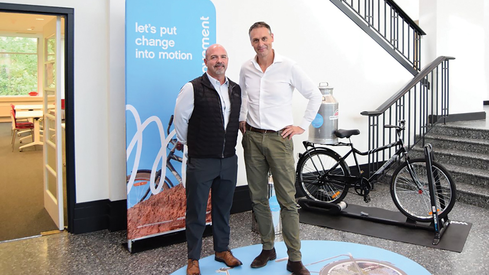 David Major and one other person standing in front of a buffalo bicycle, in a building with white walls and black floors.