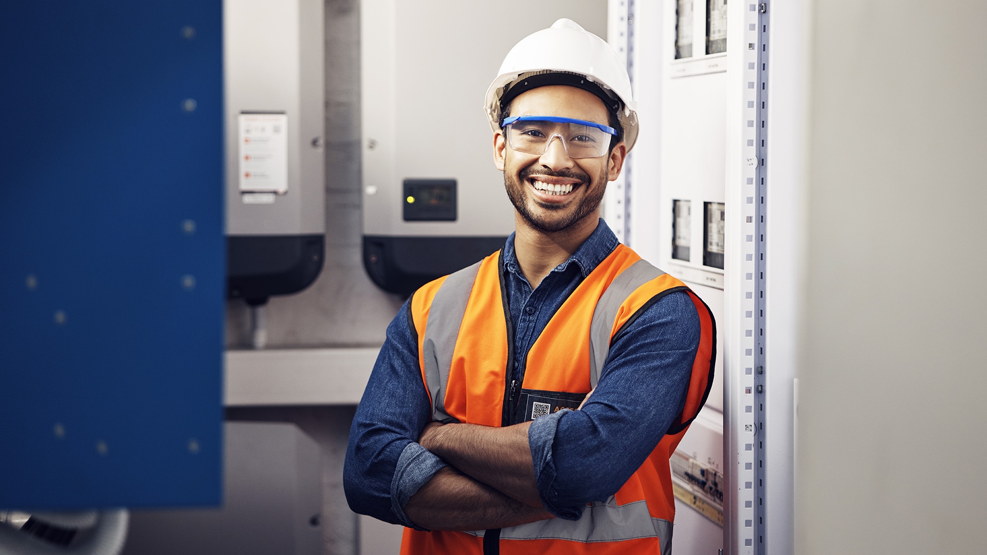 A man in an orange high visibility vest standing with his arms crossed in a boiler room.