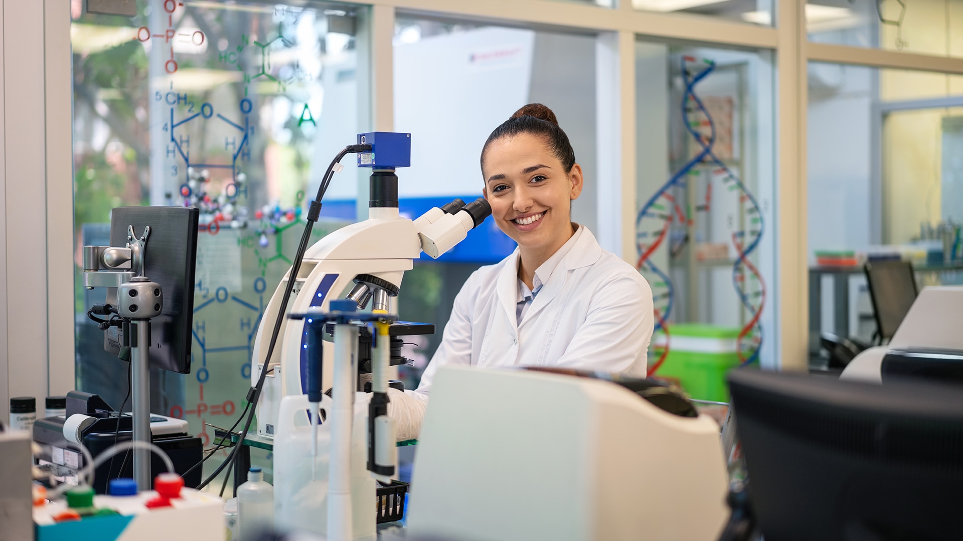 A female medical lab technician in a white lab coat standing in front of a microscope.