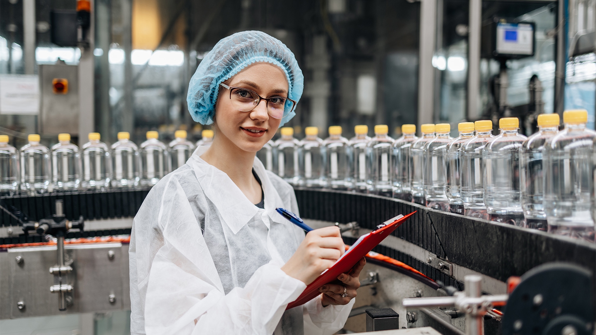 A woman in a white coat and blue hairnet standing with a clipboard in front of a conveyor belt with clear plastic bottles.