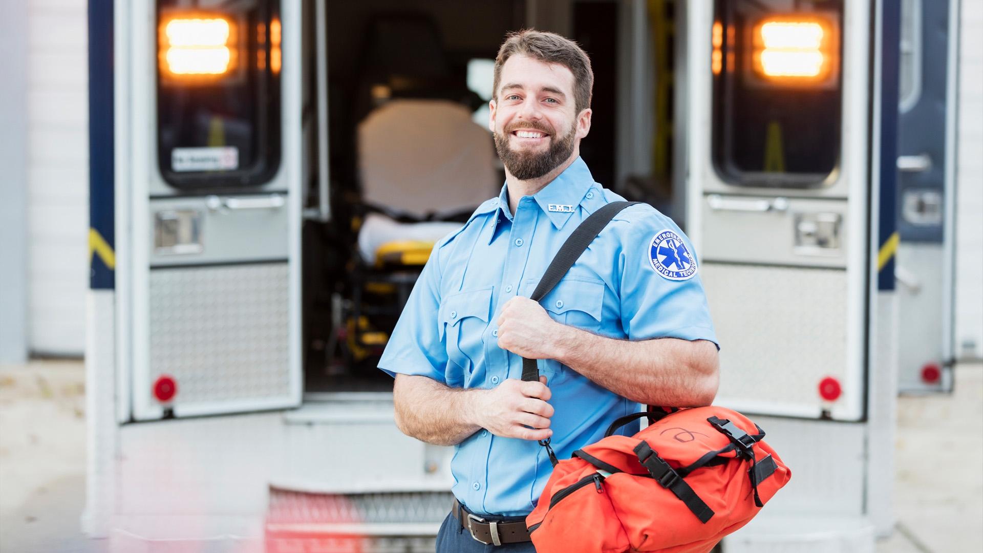 A young man in his 20s, a paramedic, standing at the rear of an ambulance, by the open doors, carrying a medical trauma bag. He is looking at the camera with a confident expression, smiling.