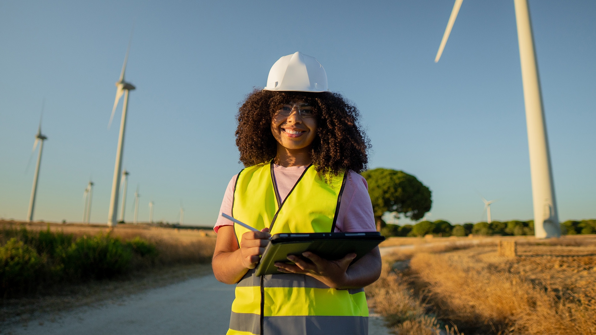 A female environmental technician in a high visibility vest and hard hat standing on a dirt road surrounded by fields.