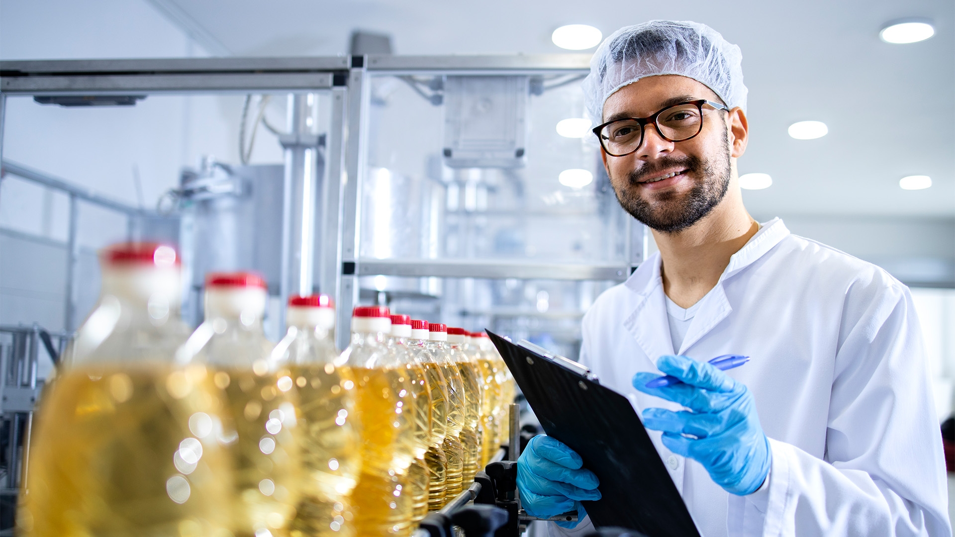 Food technician standing next to a row of oil bottles with a clipboard.