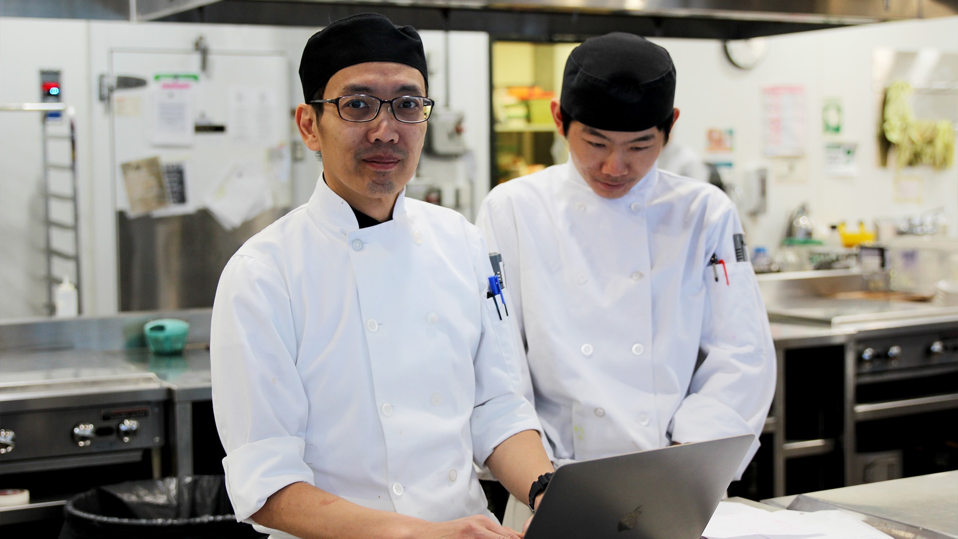 Two people standing in white uniforms in the kitchen in front of a laptop.