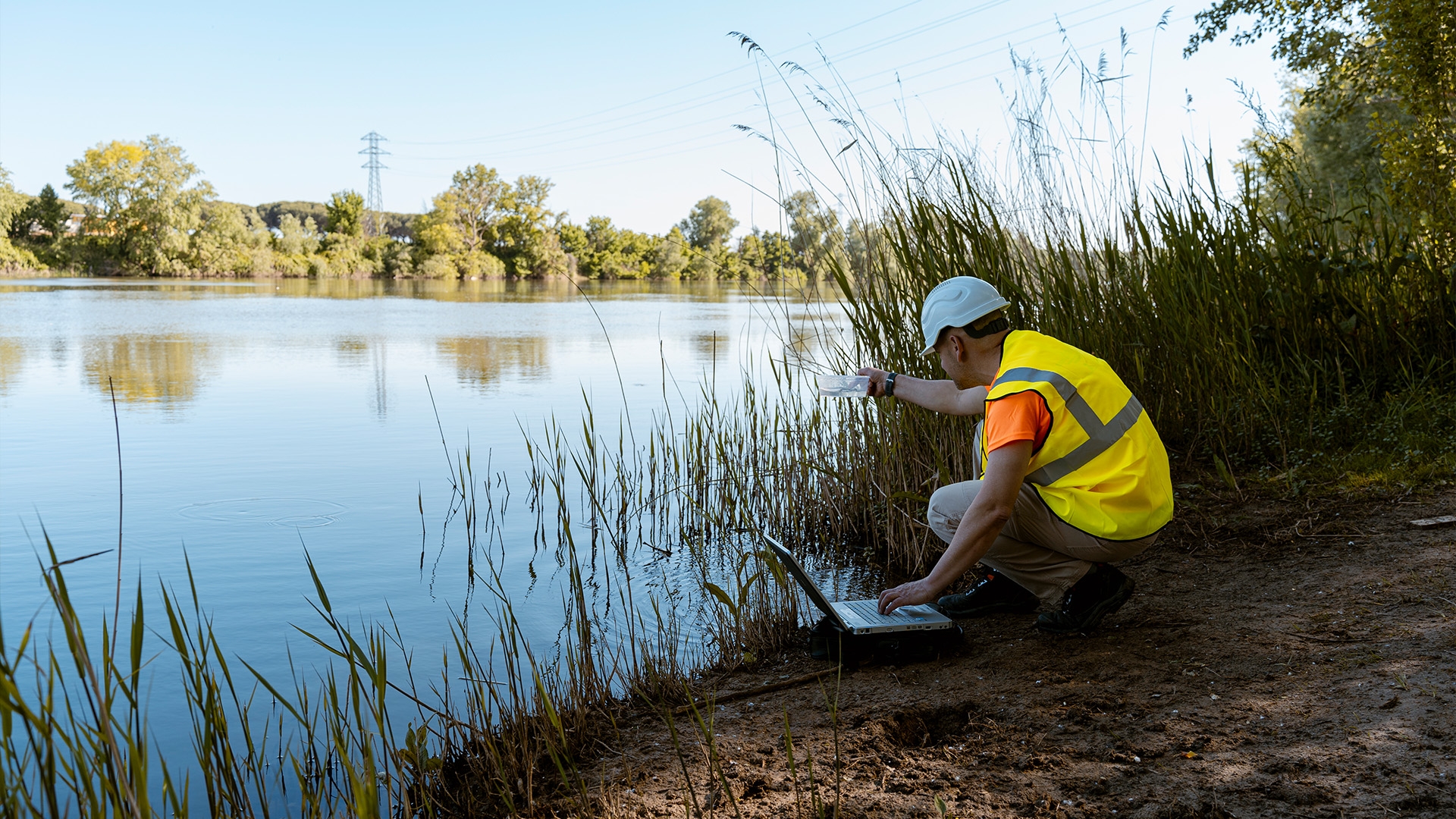 A person in a high visibility vest and hard hat crouching next to a laptop by a body of water.
