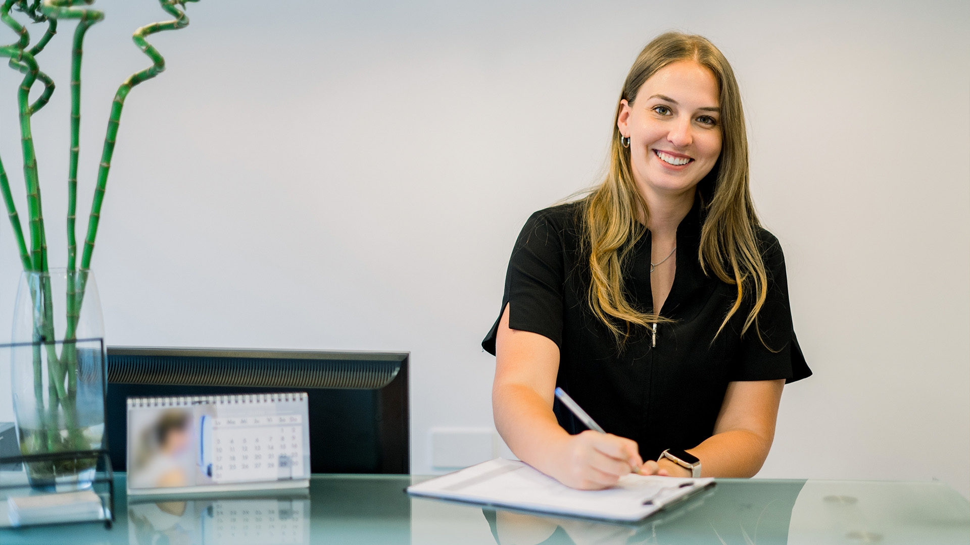 A smiling receptionist, dressed in a black uniform, sits behind a sleek glass desk, greeting patients at an aesthetic clinic. She holds a clipboard and pen, ready to assist with patient forms.