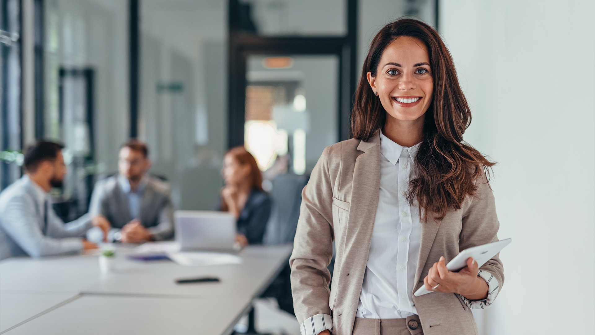 A confident professional woman stands in a modern office holding a tablet, while in the background, a diverse team of colleagues collaborate at a table.