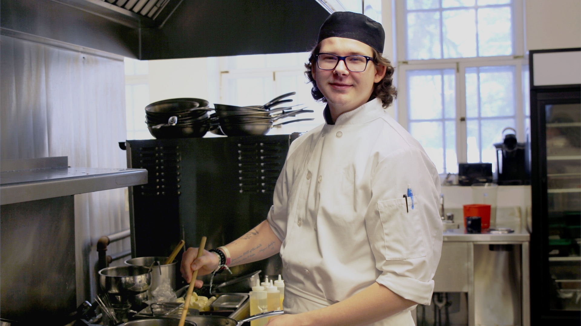 A student in a white uniform standing in front of a stove, stirring something in a saucepan while looking at the camera.