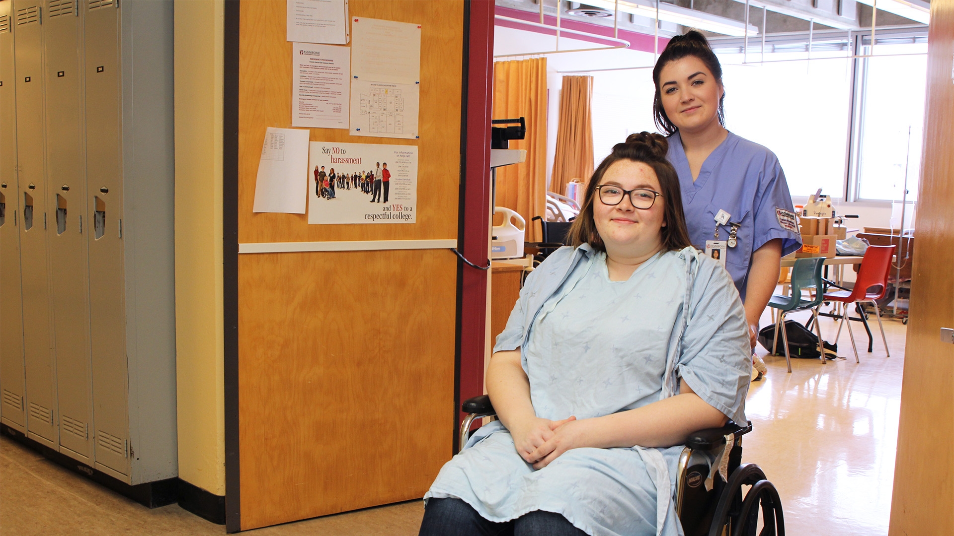 A student in blue scrubs standing behind a person in a wheelchair at an entrance to a classroom.
