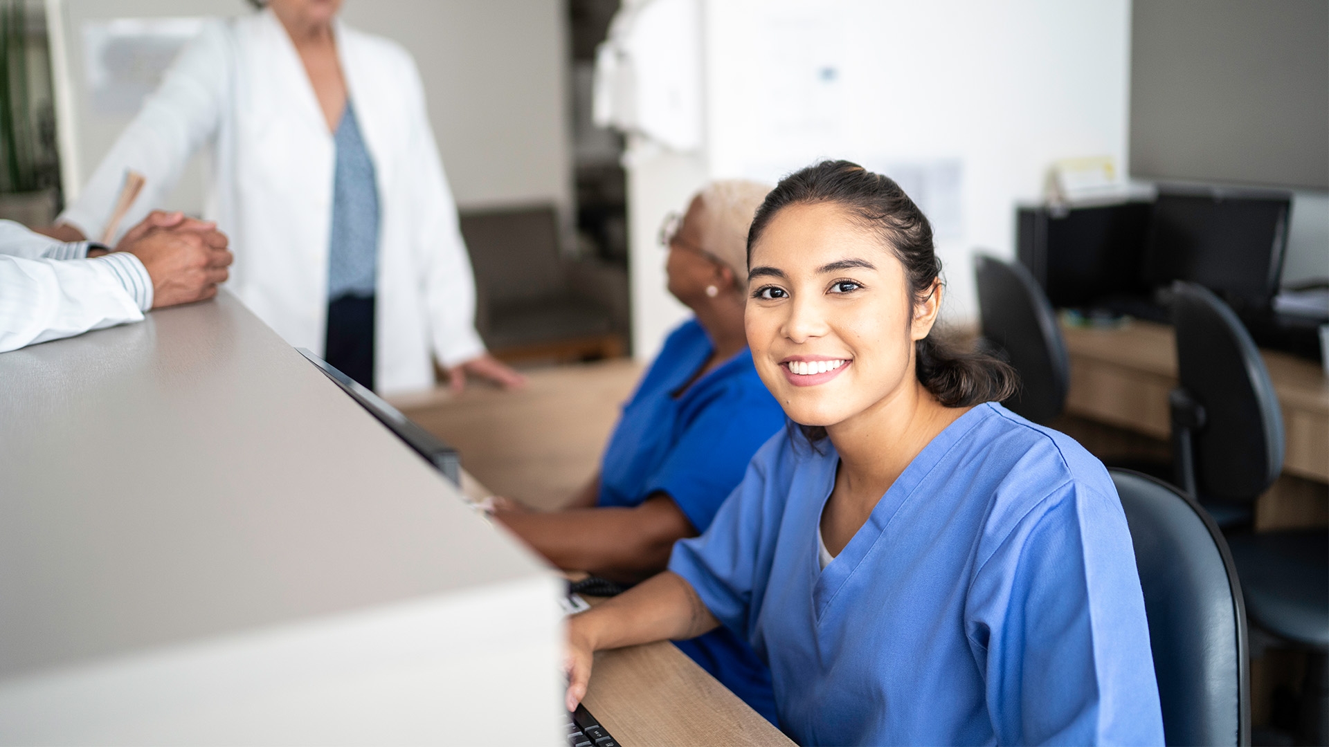 A medical administration assistant sitting at the front desk of a medical office in blue scrubs.