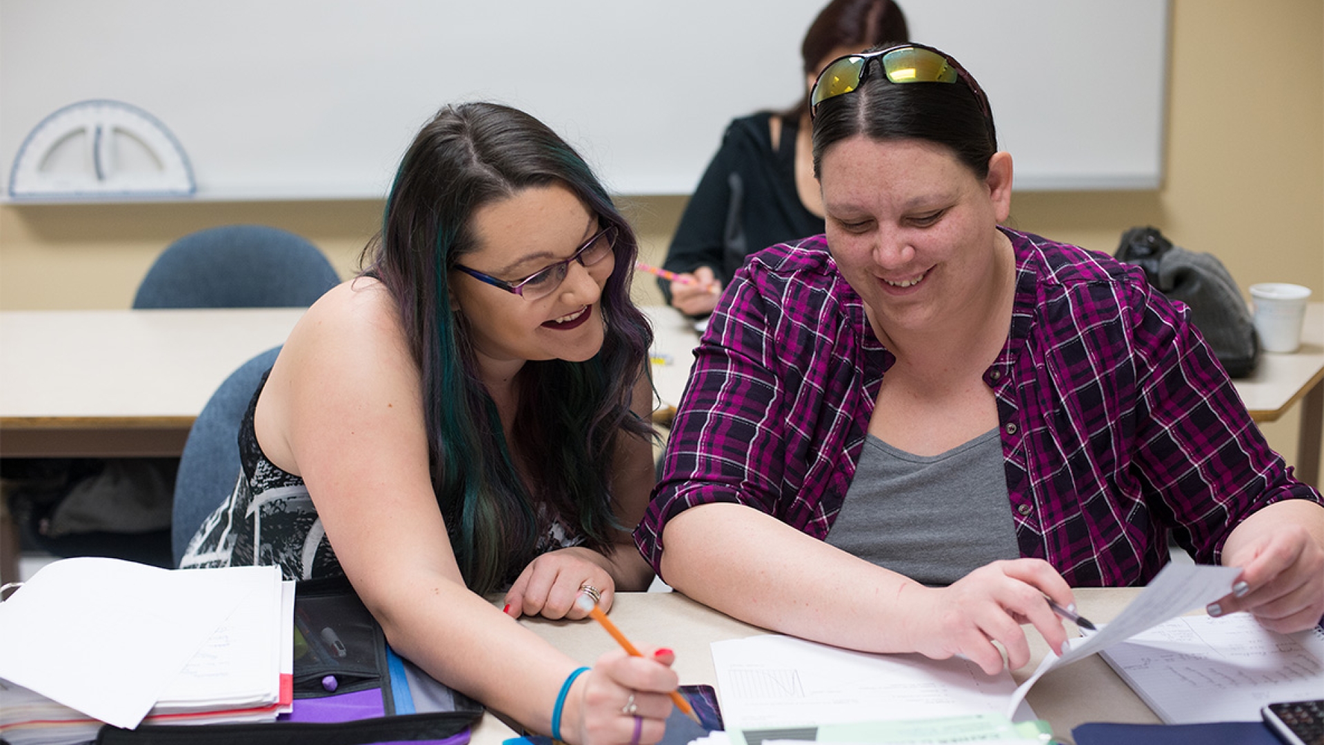 Two students sitting in a classroom, looking at a textbook together.