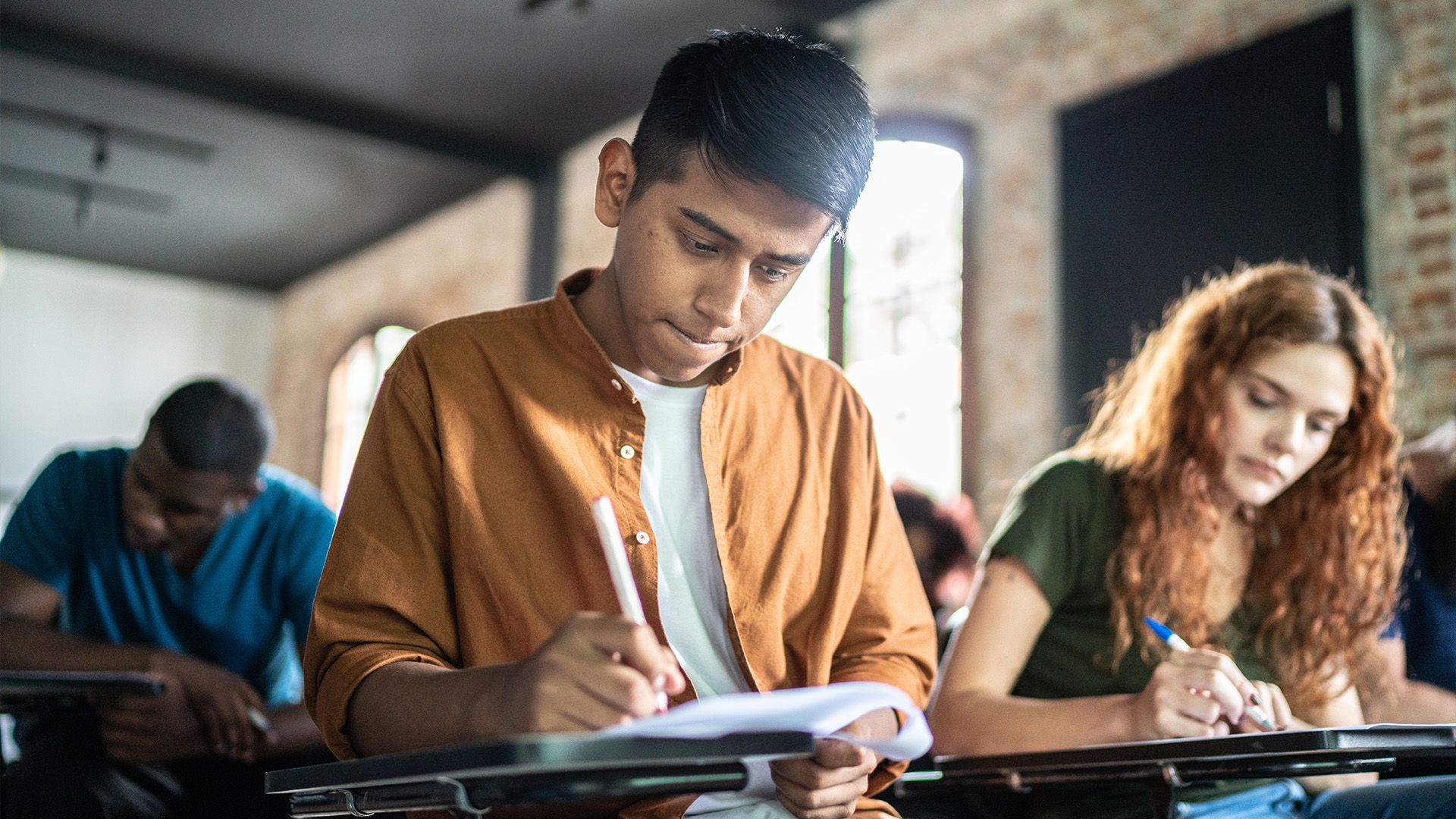 A male student in an orange shirt writing on paper during class.