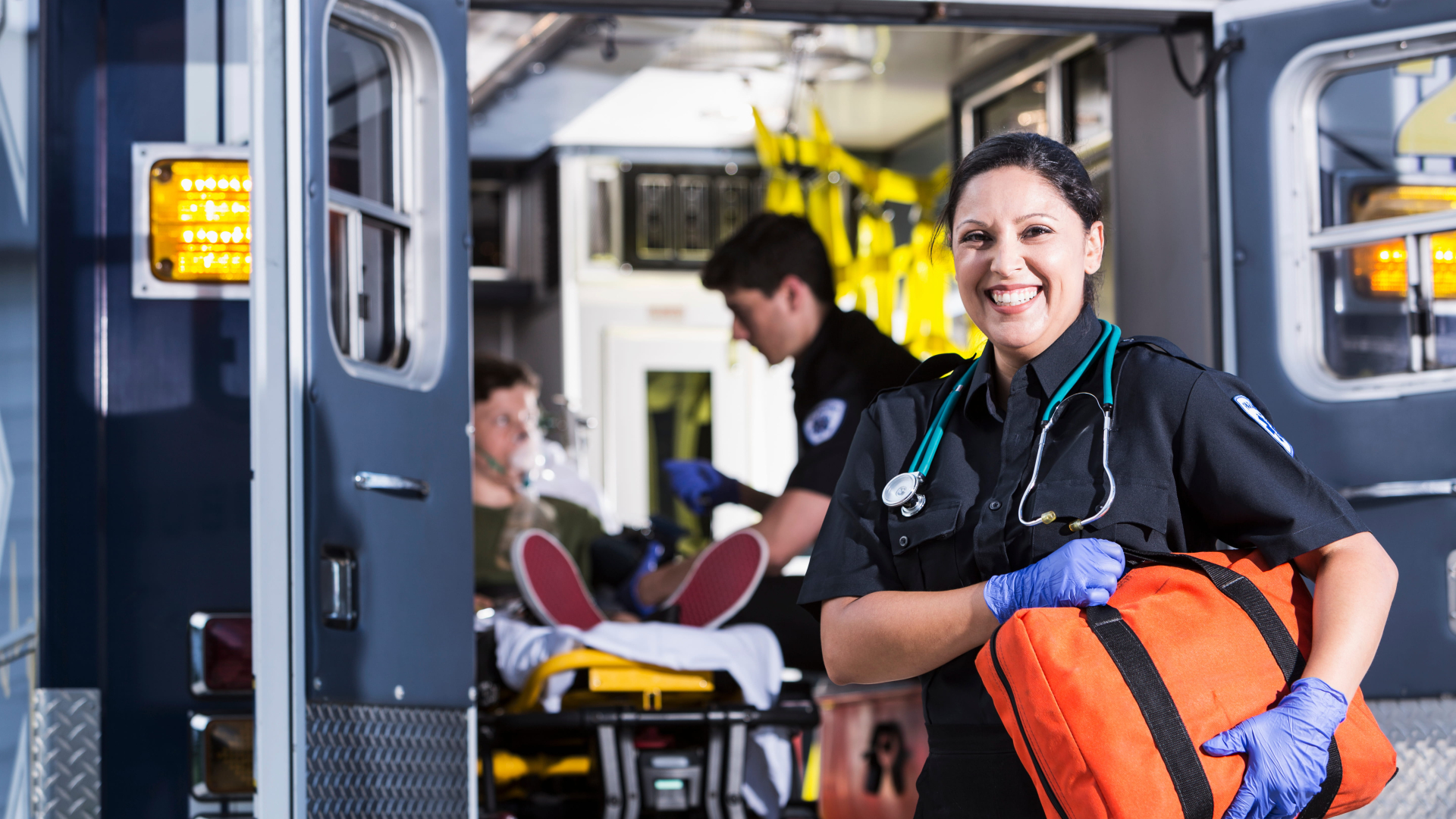 lady in her 30's holding a orange bag and standing in font of the back of an ambulance with the doors open an a patient sitting on a stretcher with a oxygen mask, being assisted by a male paramedic