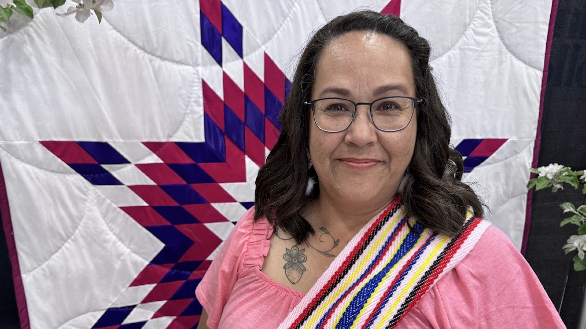 Tammy Chief wearing a pink shirt and a Métis sash, standing in front of the Assiniboine quilt.