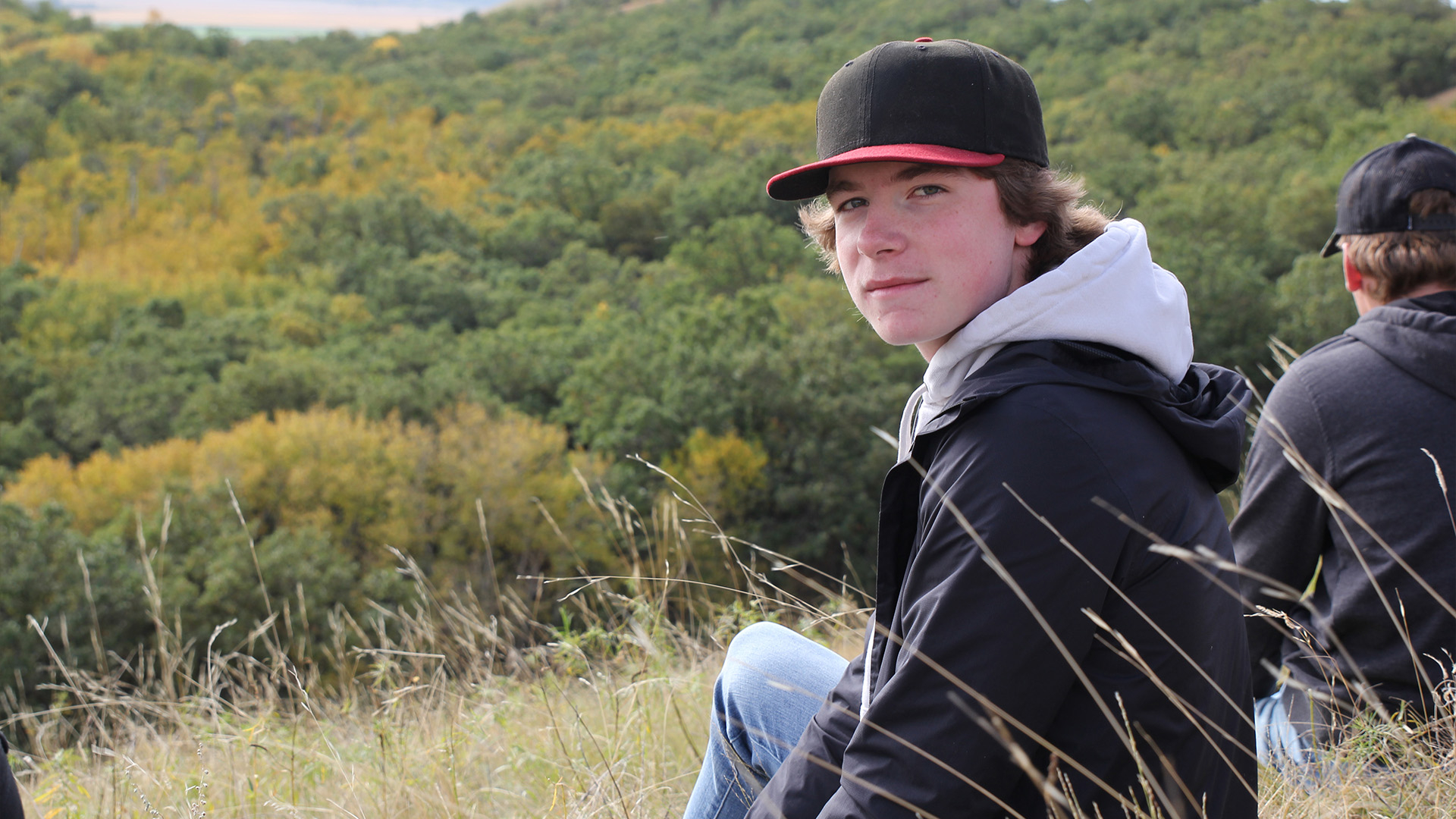 A male student in a cap and black jacket sitting on the side of a hill.