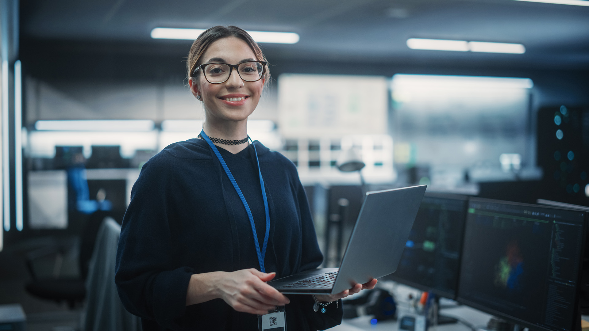 A woman wearing a black sweater and glasses, standing with a laptop in a room full of computers and server racks.