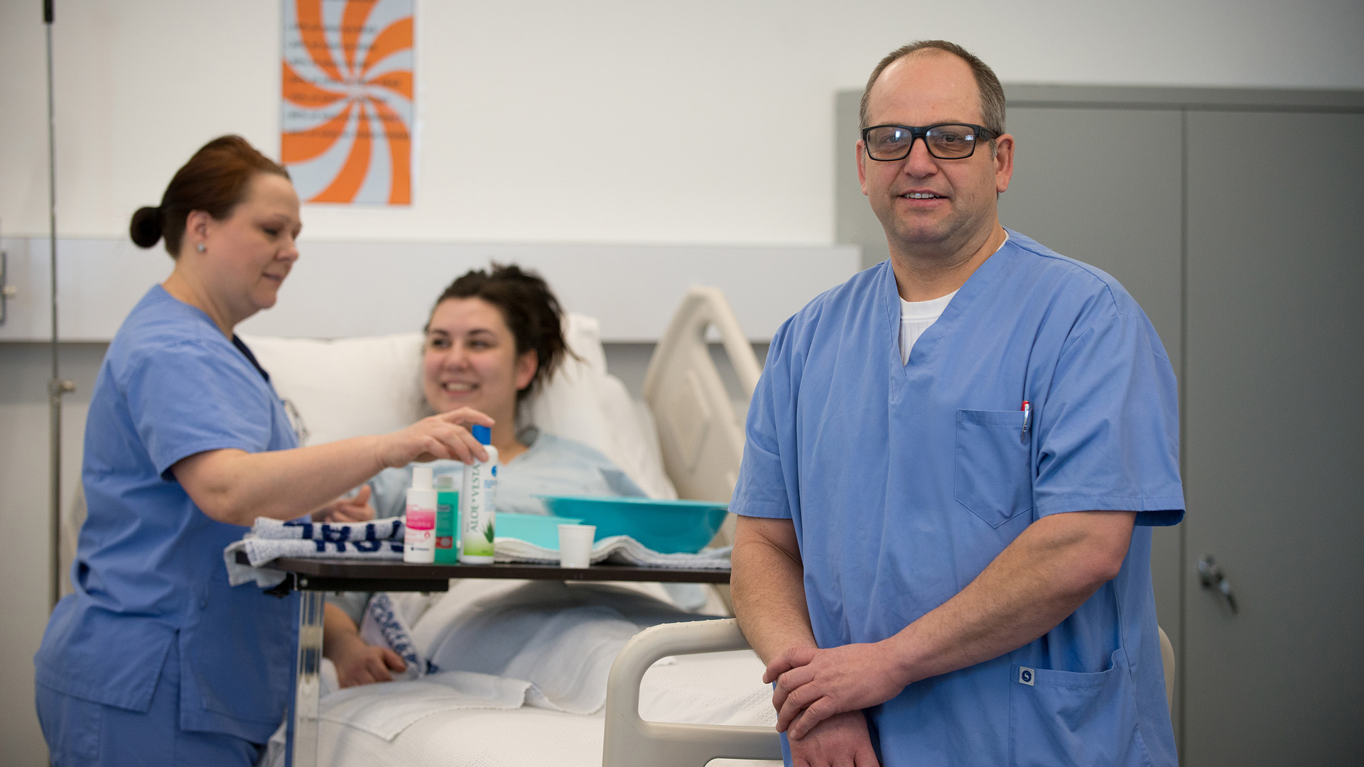 A health care aide in blue scrubs posing for a photo, while another aide is talking to a patient in a bed behind him.