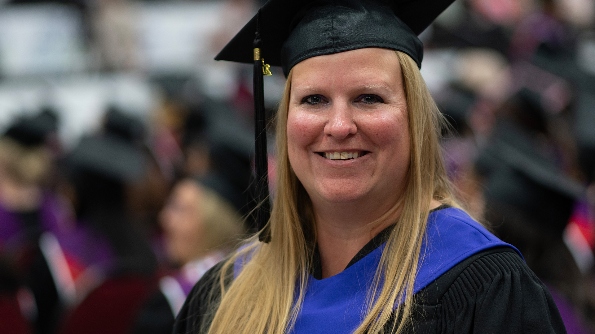 A graduate with long blond hair, a black cap and gown with a blue hood standing in a crowd of other graduates.