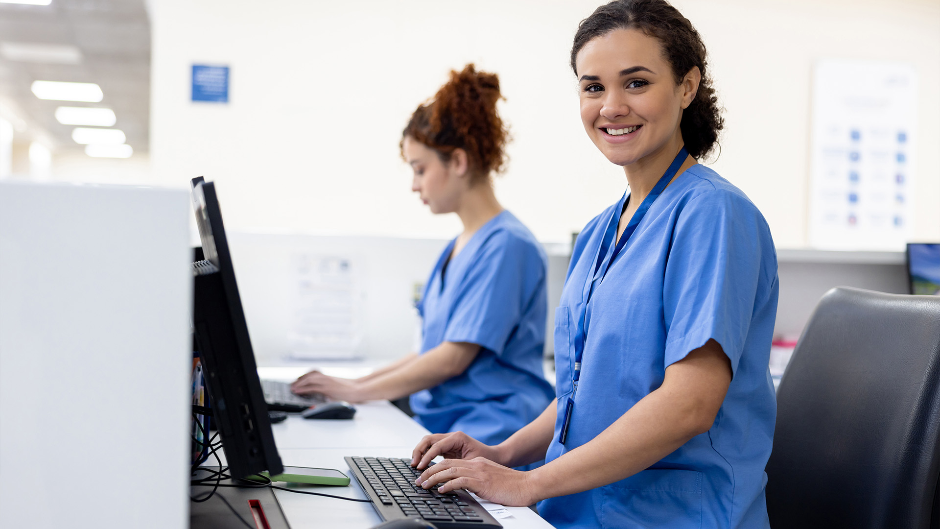 Two women in blue scrubs standing at the reception desk, typing information into the computers.